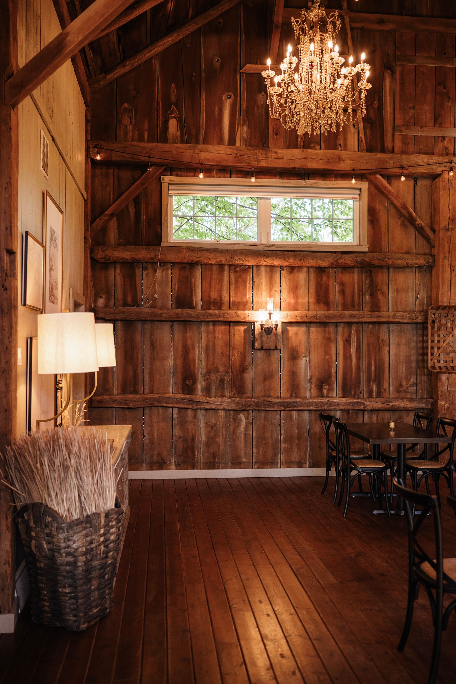 Rustic barn interior with wood walls, crystal chandelier, wheat basket, and dining chairs bathed in warm golden light.