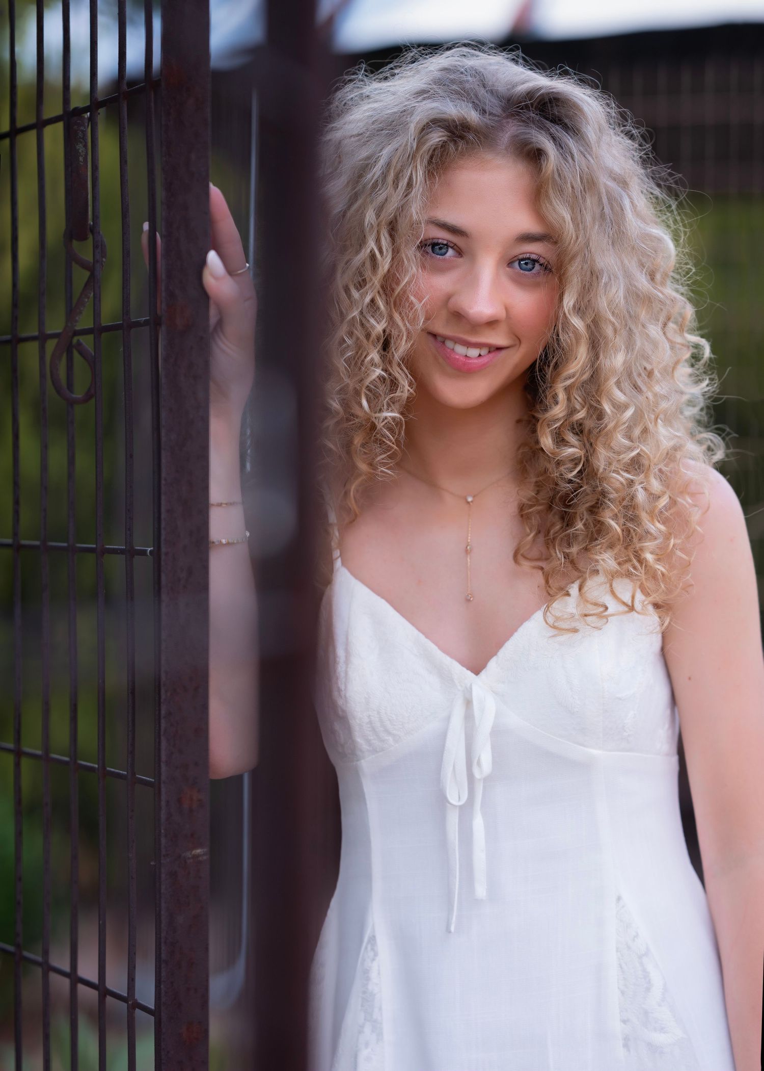 Young woman with curly blonde hair smiling in a white sundress, leaning against a rustic metal gate outdoors.