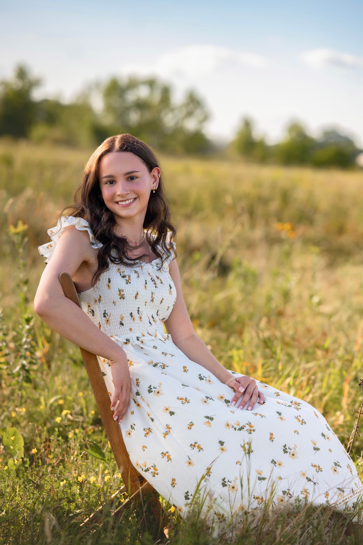 Young woman in floral white dress sitting in a sunlit meadow, smiling warmly during golden hour.
