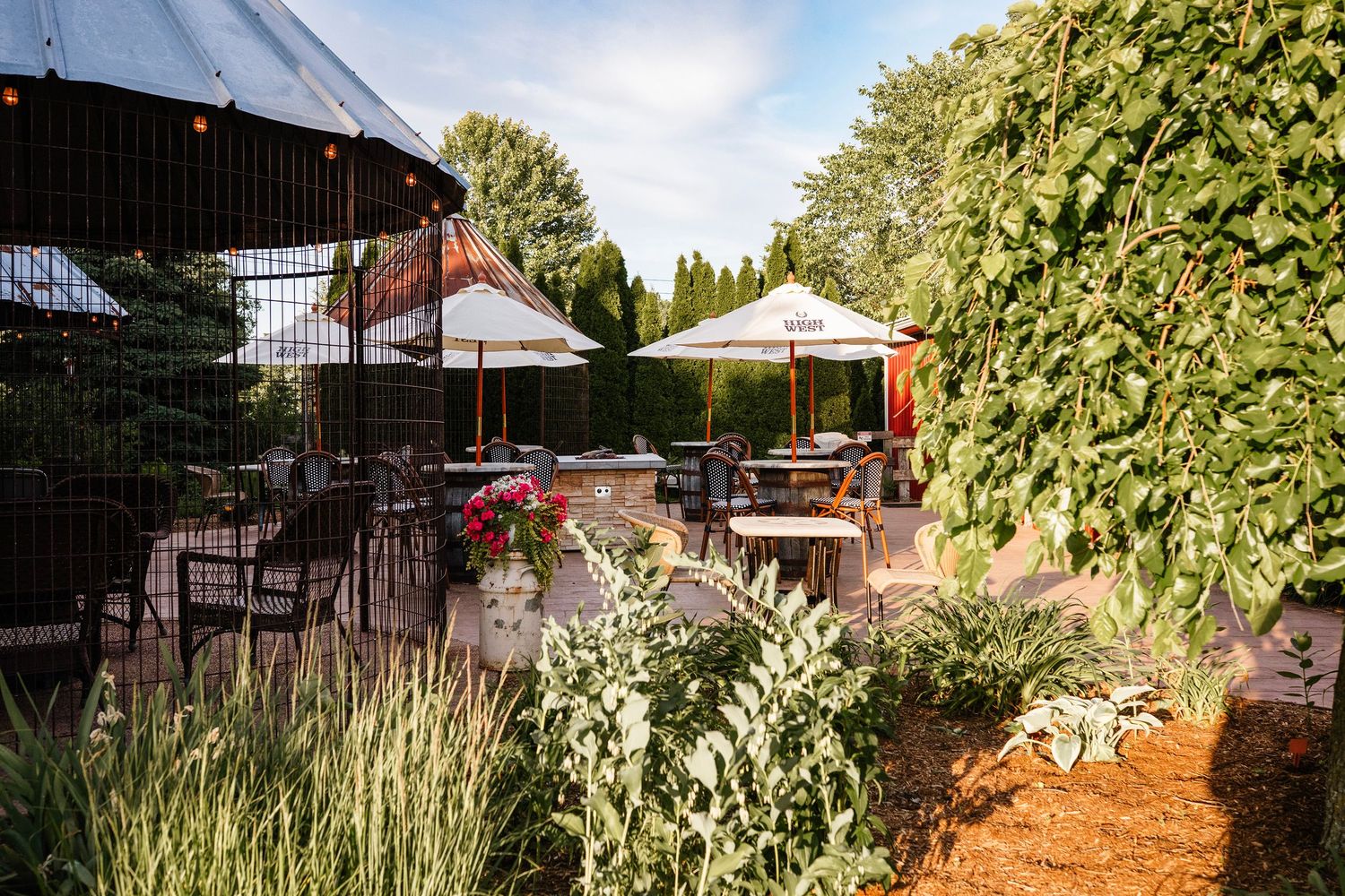 Outdoor restaurant patio with white umbrellas, dining tables, lush greenery, and garden landscaping on a sunny day.