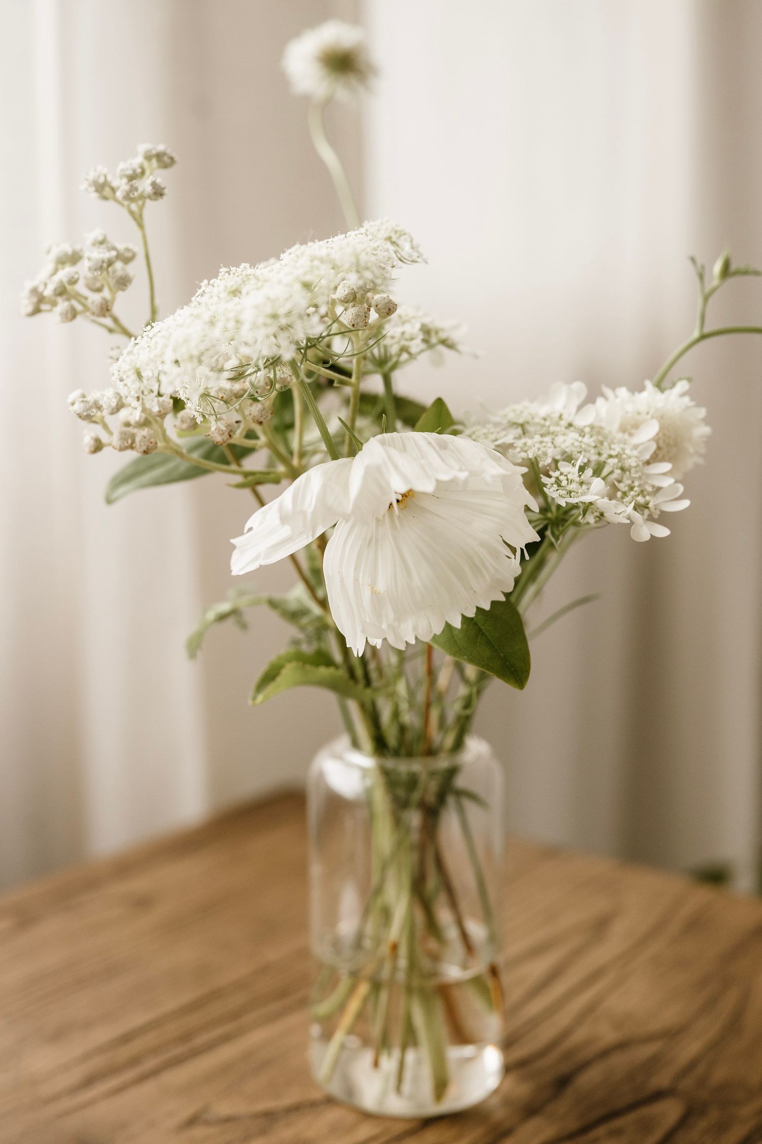 White wildflowers and ranunculus in a clear glass jar vase on a rustic wooden table with soft natural light.