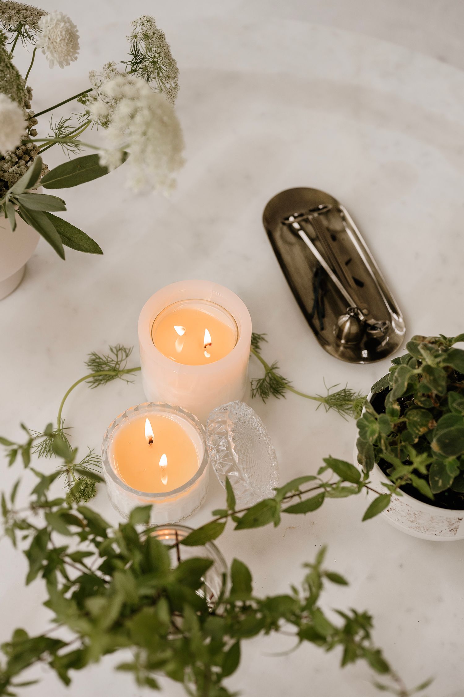 Two lit pillar candles surrounded by green foliage and white flowers on a marble surface with a silver decorative object.