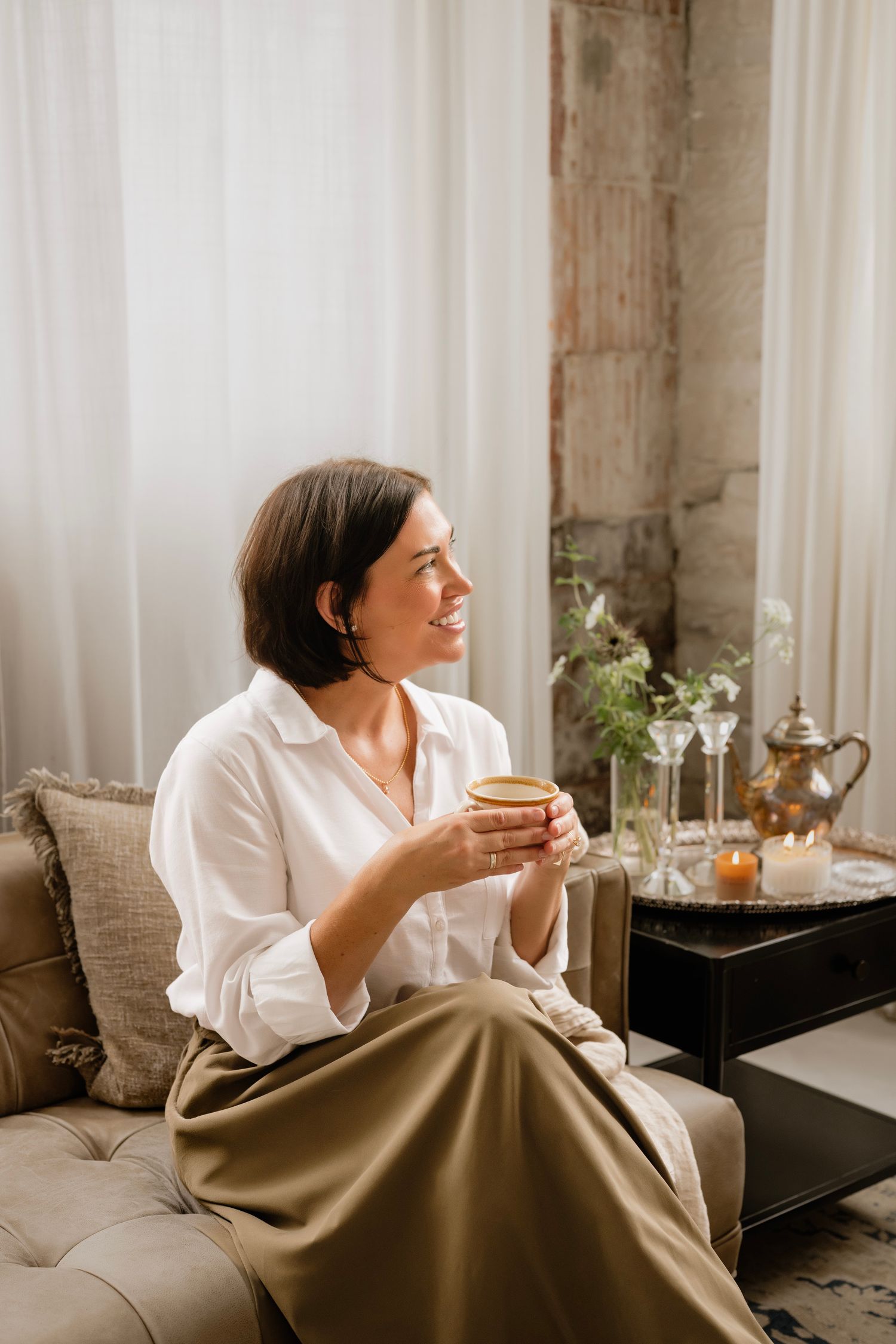 Smiling woman in white blouse holding coffee cup, sitting on beige sofa in elegant living room.