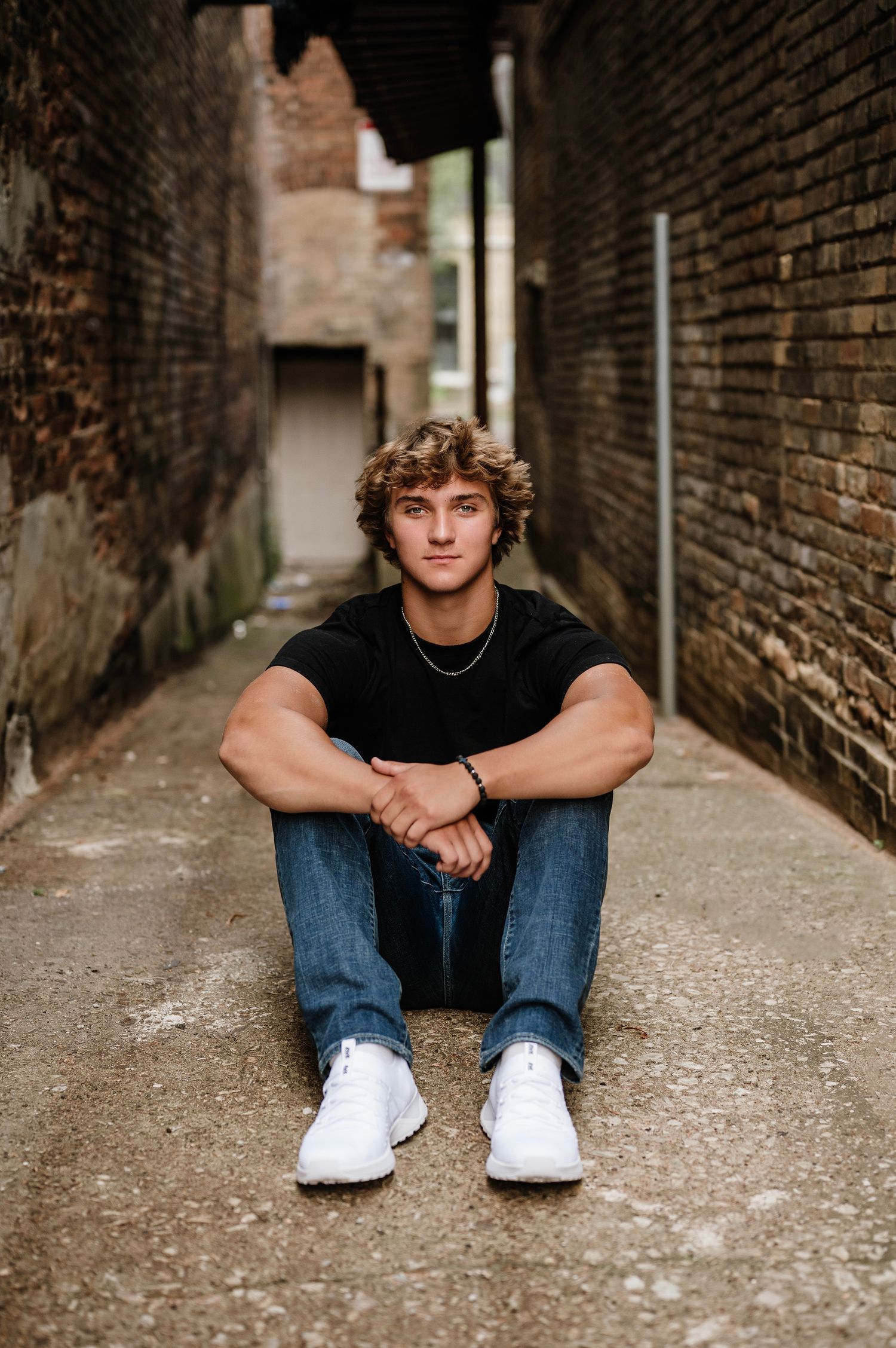 A young person in casual attire sits in a brick alleyway wearing jeans and white sneakers in a moody outdoor portrait series.