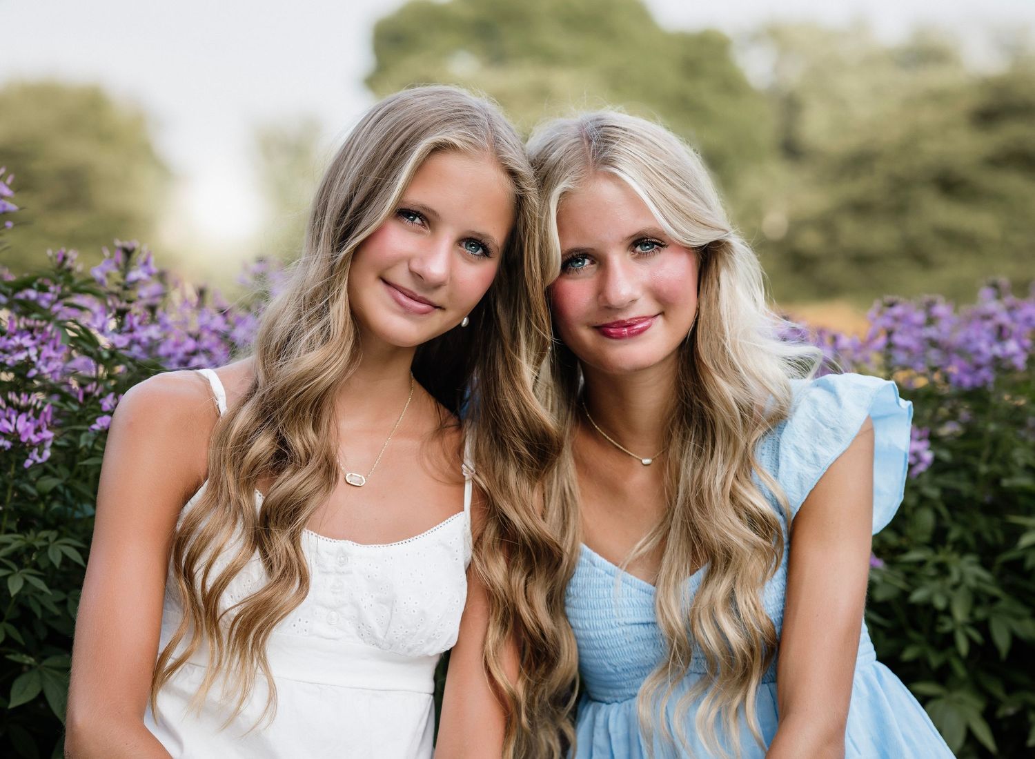 Two people in white and blue dresses pose together in a garden with purple flowers in the background.