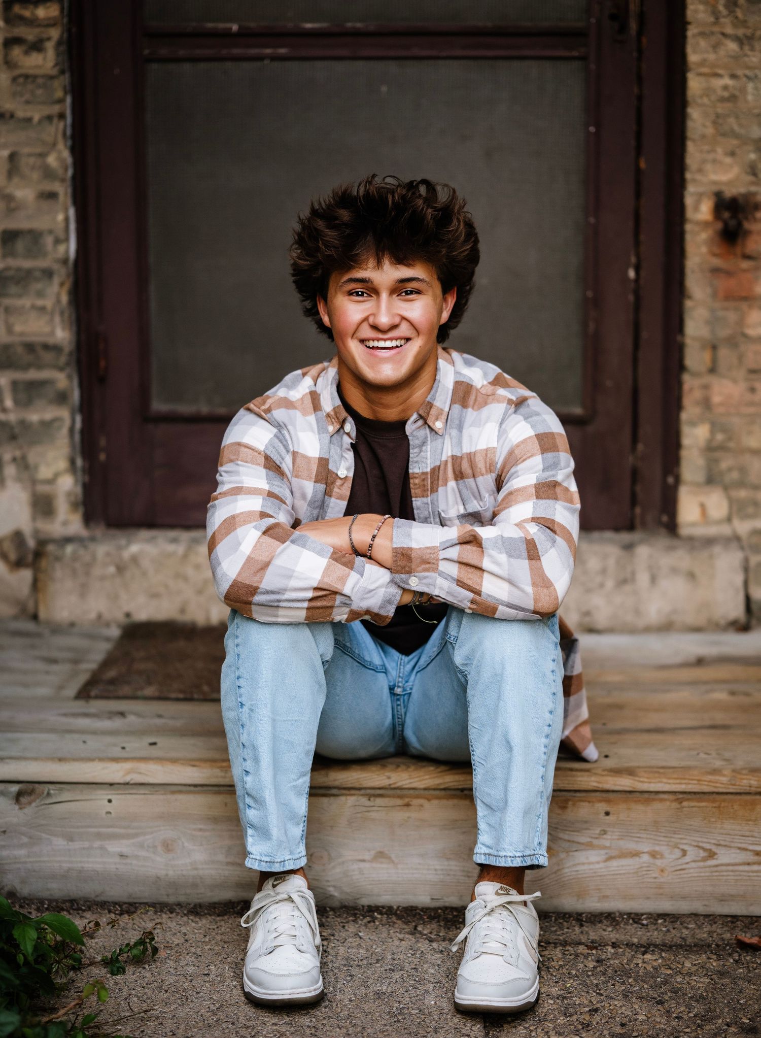 Person in plaid shirt and jeans sits casually on concrete steps in front of brick building.