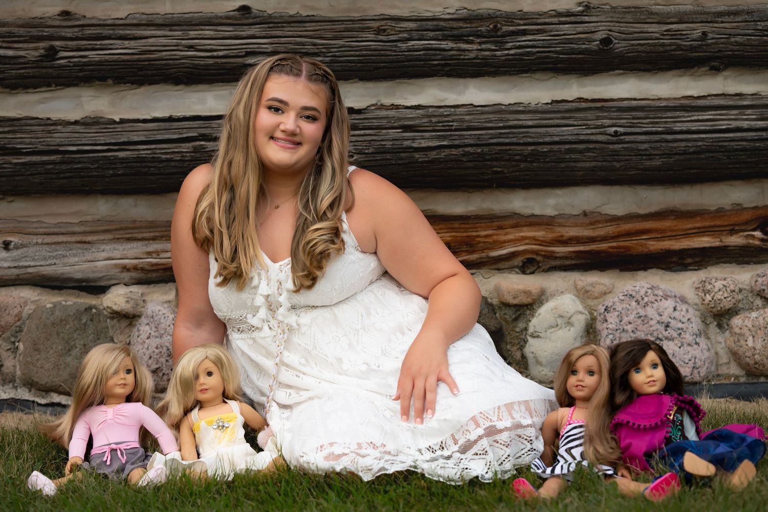 Teen girl in white dress sitting on grass with American Girl dolls in front of a rustic log wall.