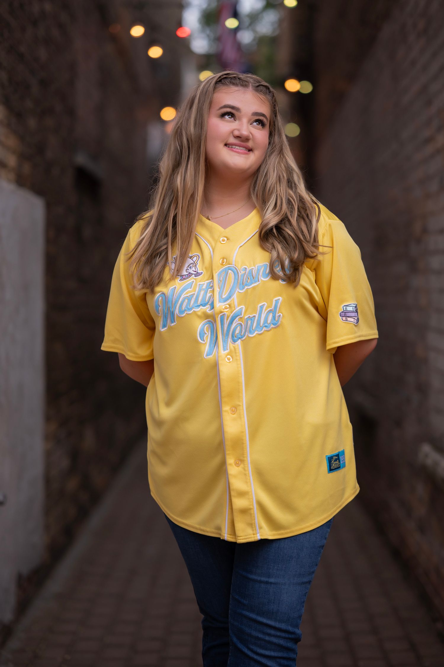 Young woman smiling in yellow Walt Disney World baseball jersey, posing in brick alleyway with bokeh lights.