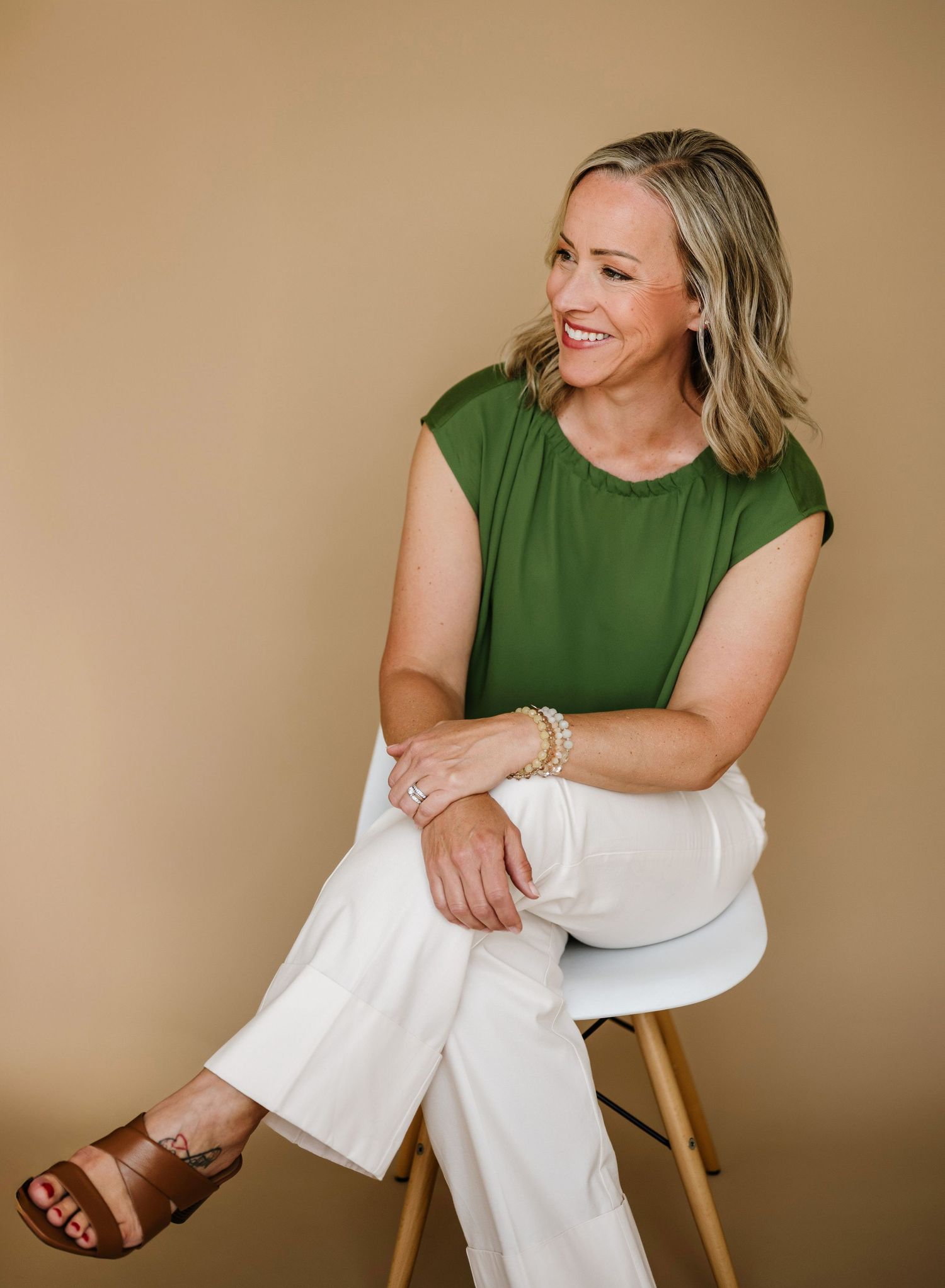 A woman in a green sleeveless top and white pants sits casually on a wooden chair against a beige background.
