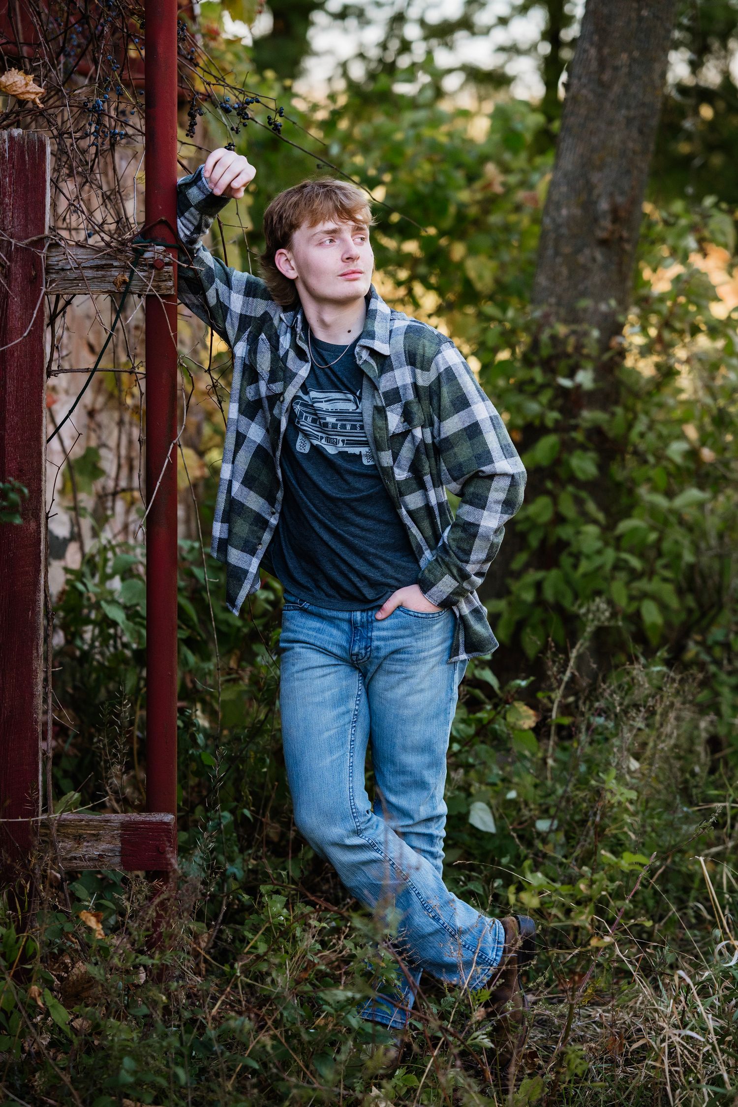A person in overalls and plaid shirt poses casually against red metal posts in an outdoor garden setting.