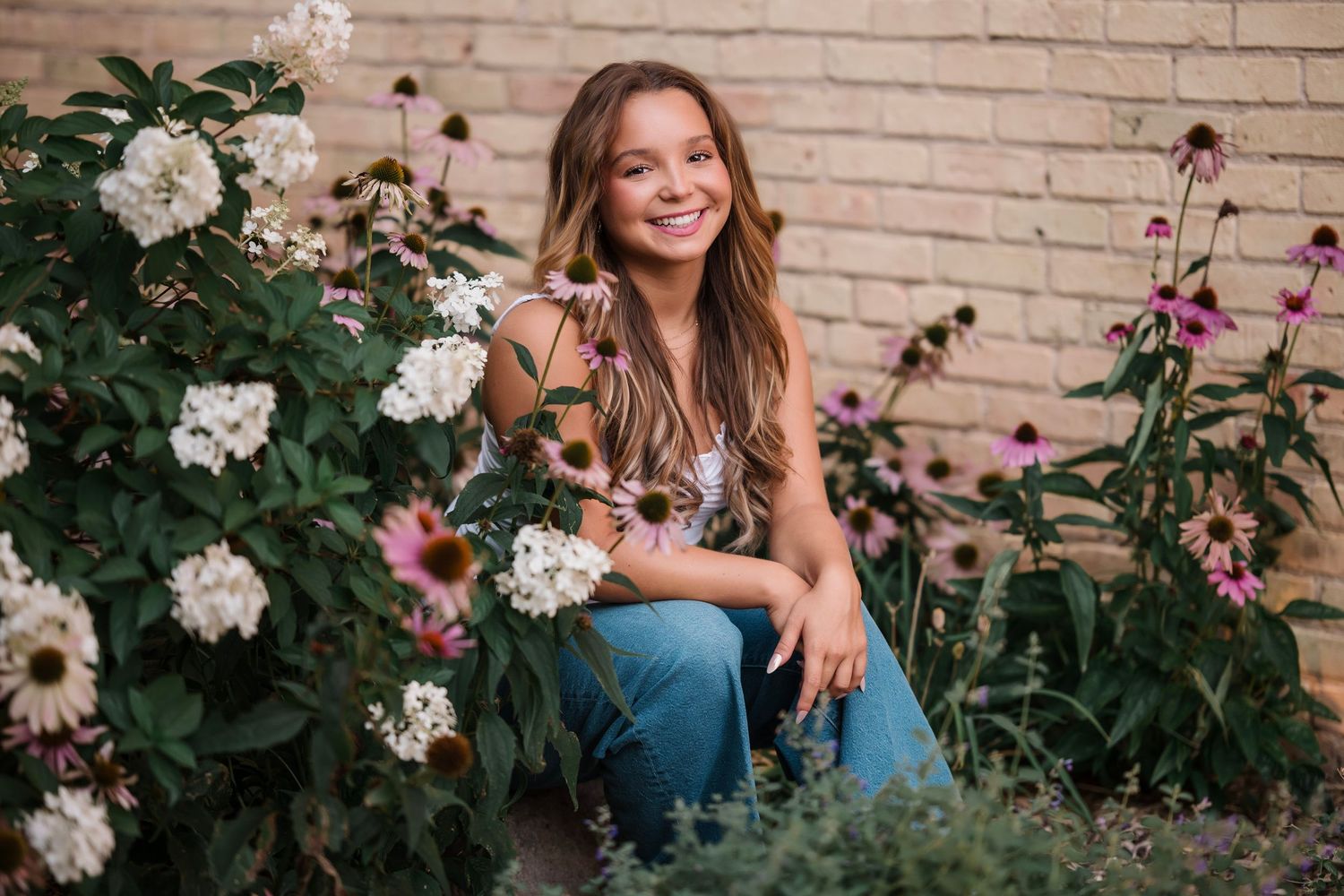 Smiling young woman with long brown hair sitting among blooming white and pink coneflowers in a garden beside a brick wall.