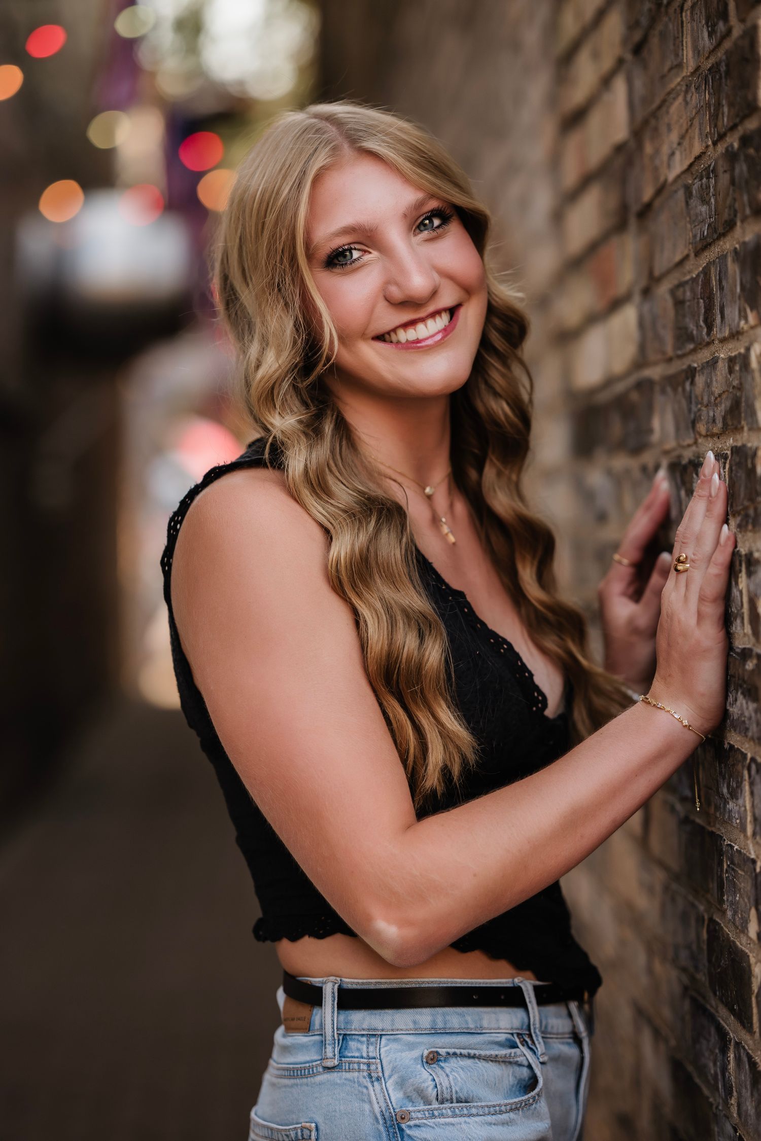 Smiling blonde woman in black crop top and jeans leaning against brick wall in urban alley setting.
