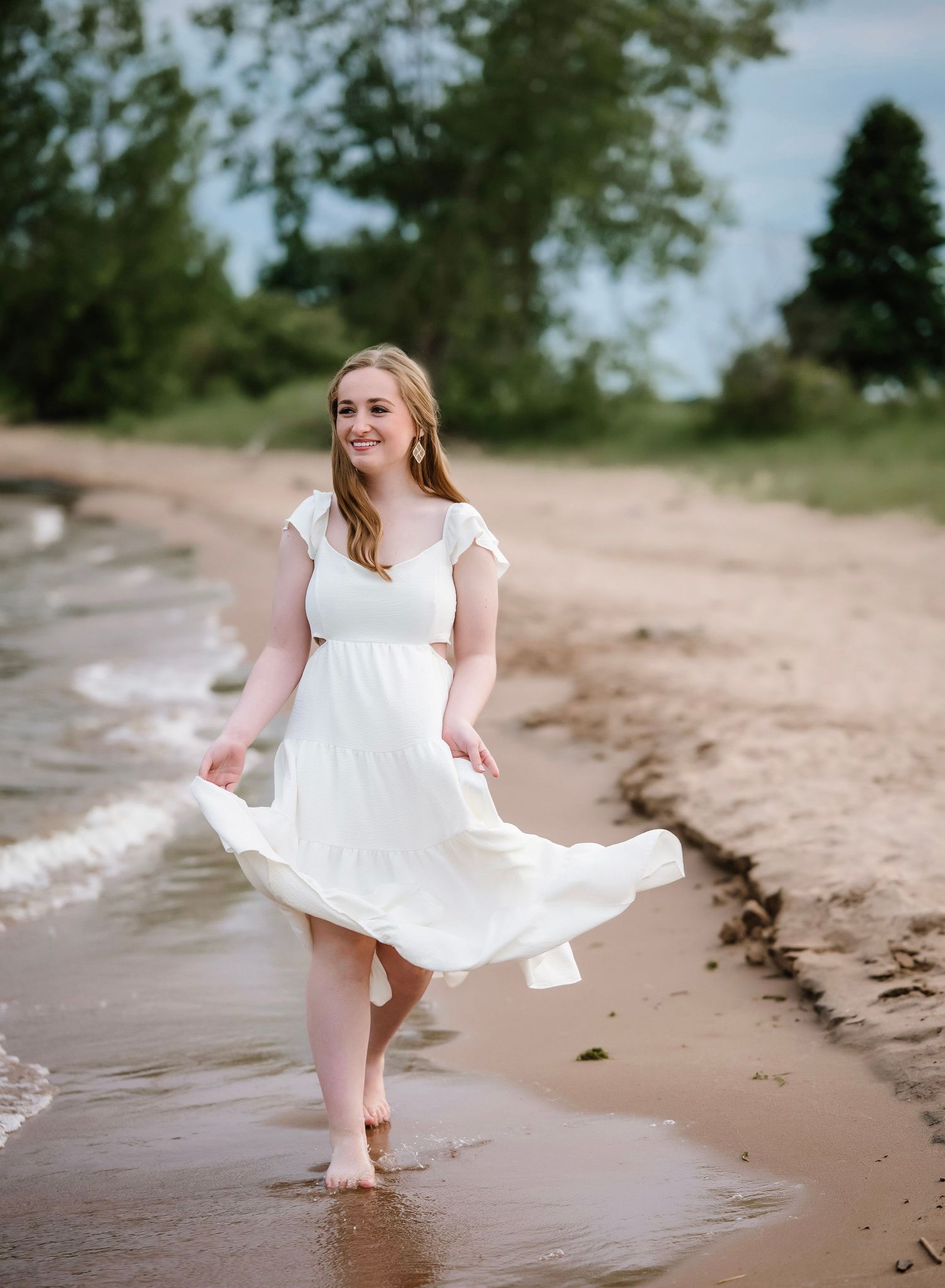 Young woman in white flowing dress walking barefoot along sandy beach shoreline with trees in background.