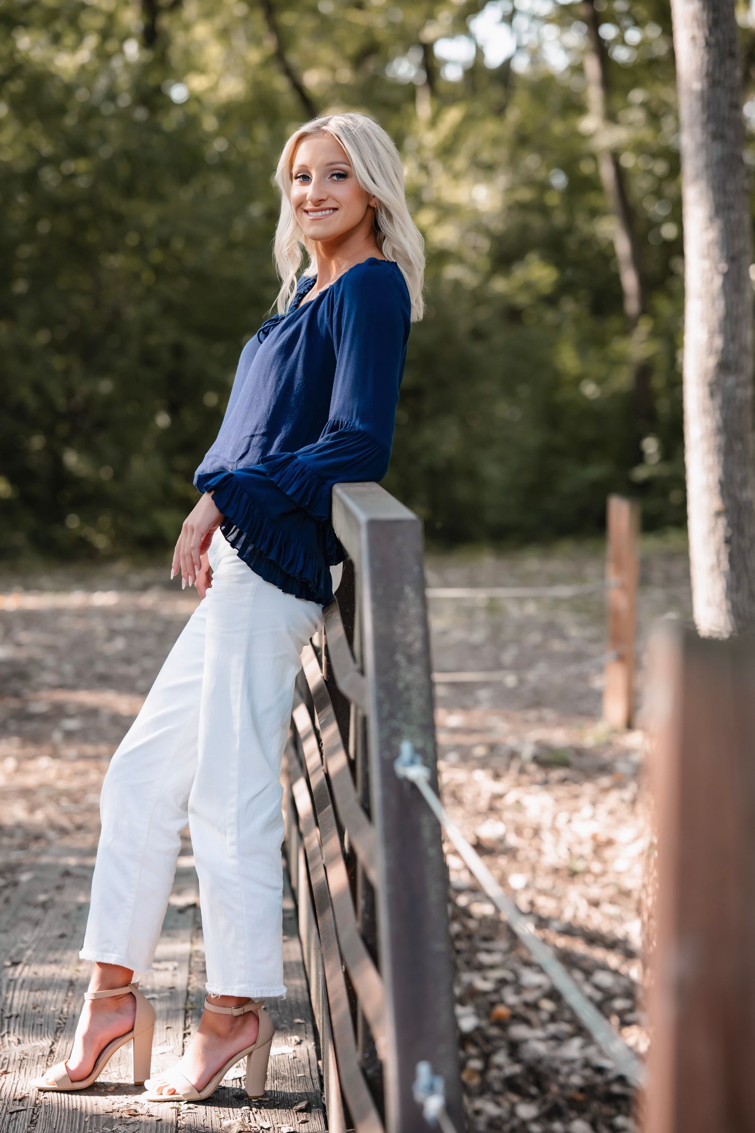 A person in a navy blue top and white pants poses outdoors near a wooden fence in a natural setting.