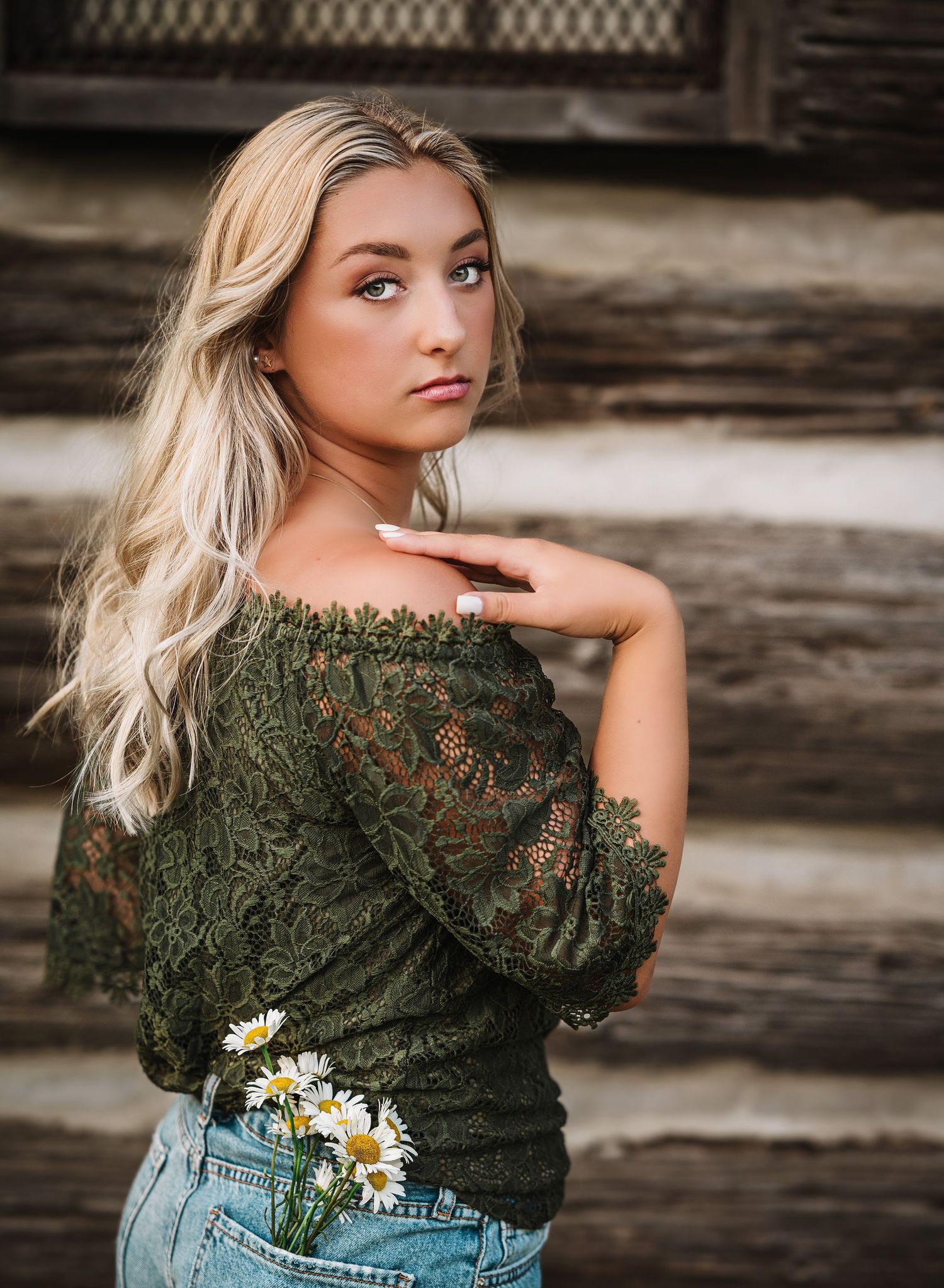 Portrait in nature with olive green lace top, light denim, and delicate daisies tucked in back pocket, against rustic wooden backdrop.