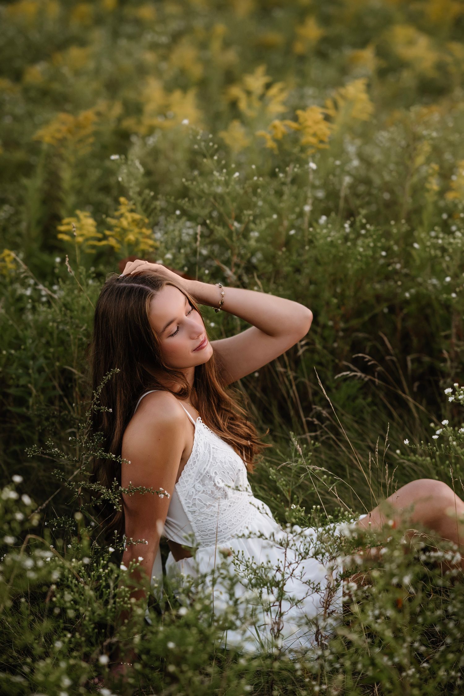 Young woman in white lace dress sitting peacefully in a wildflower meadow with yellow and white blooms.