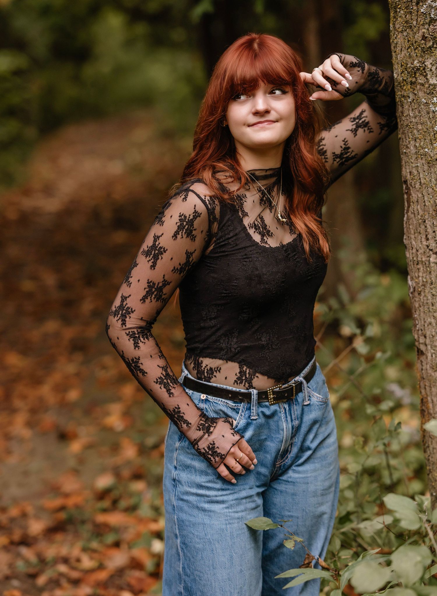 A person in a black lace top and blue jeans poses casually against a tree in an autumn forest setting.