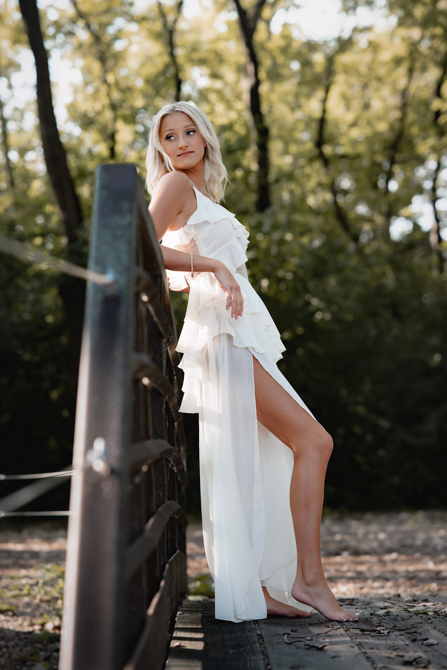 A woman in a flowing white dress poses elegantly against a natural outdoor backdrop with trees.