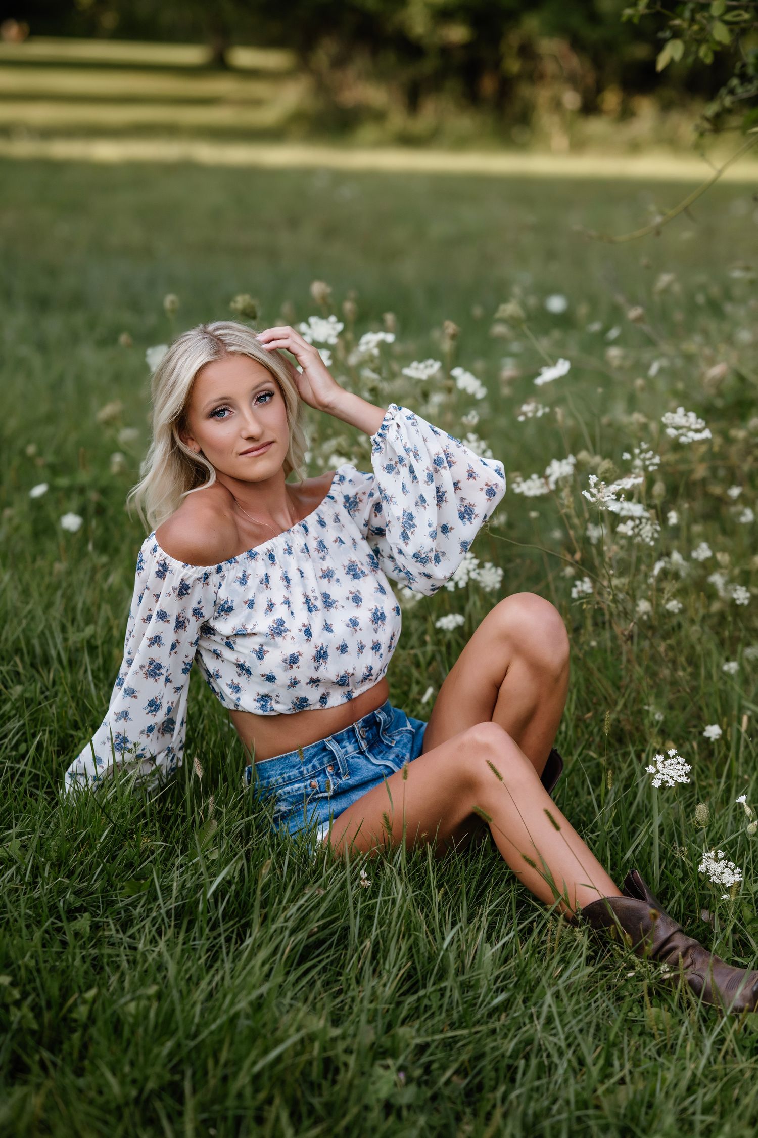 A person in a floral crop top and denim shorts reclines in a meadow of white daisies on a sunny summer day.