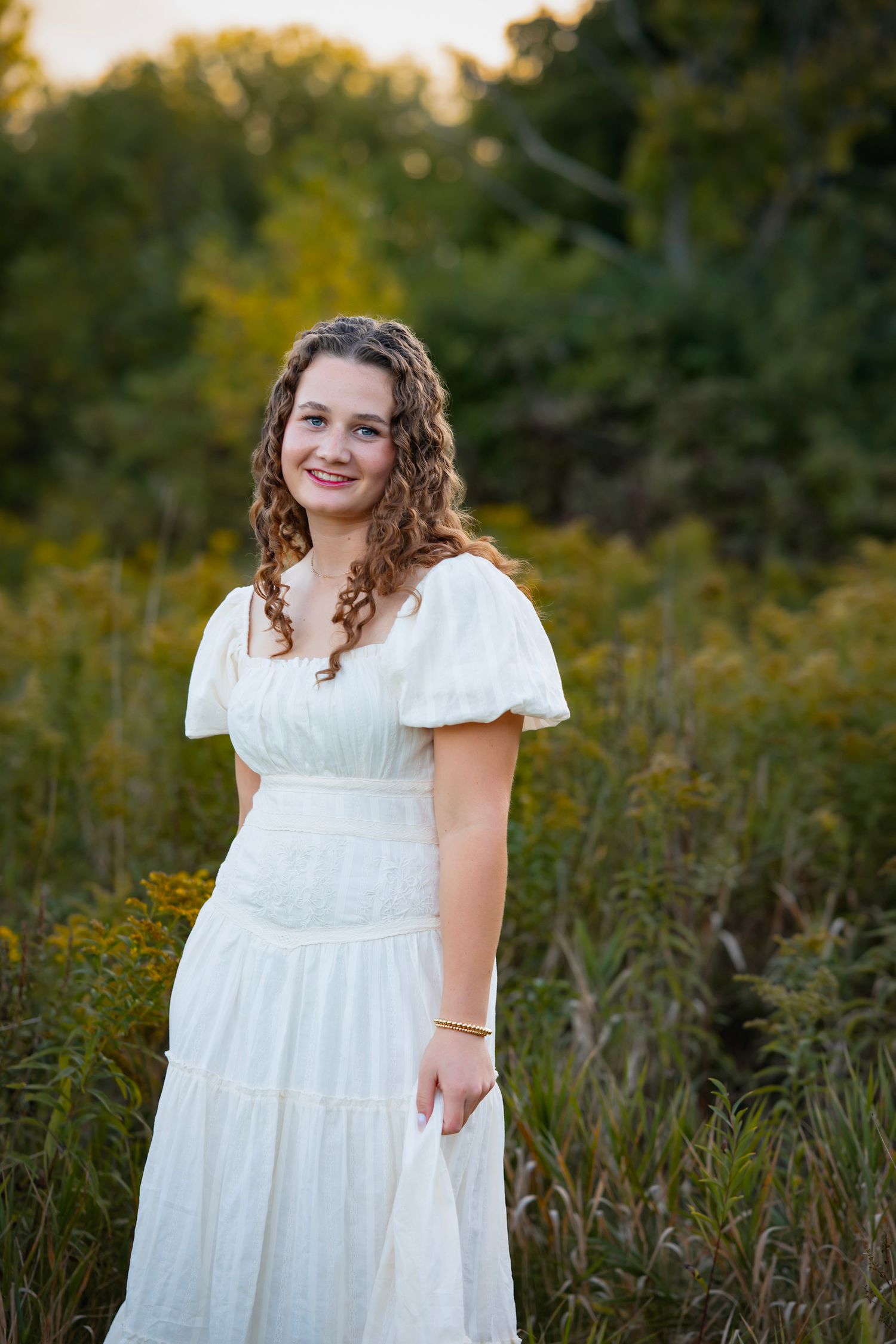 A person in a flowing white dress with puff sleeves stands in a field of tall grass at golden hour.