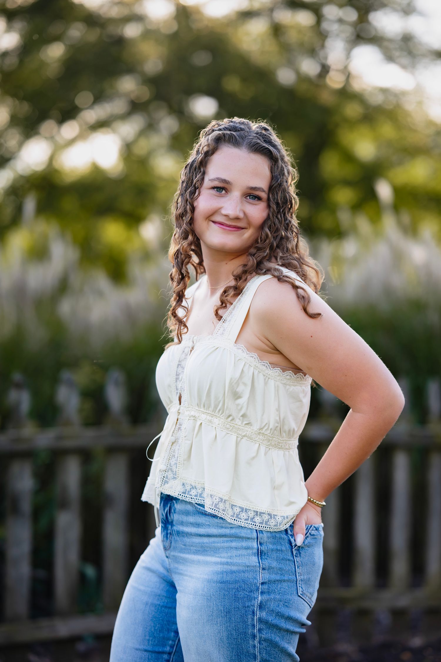 A person in a white ruffled top and blue jeans poses outdoors against a blurred natural background.