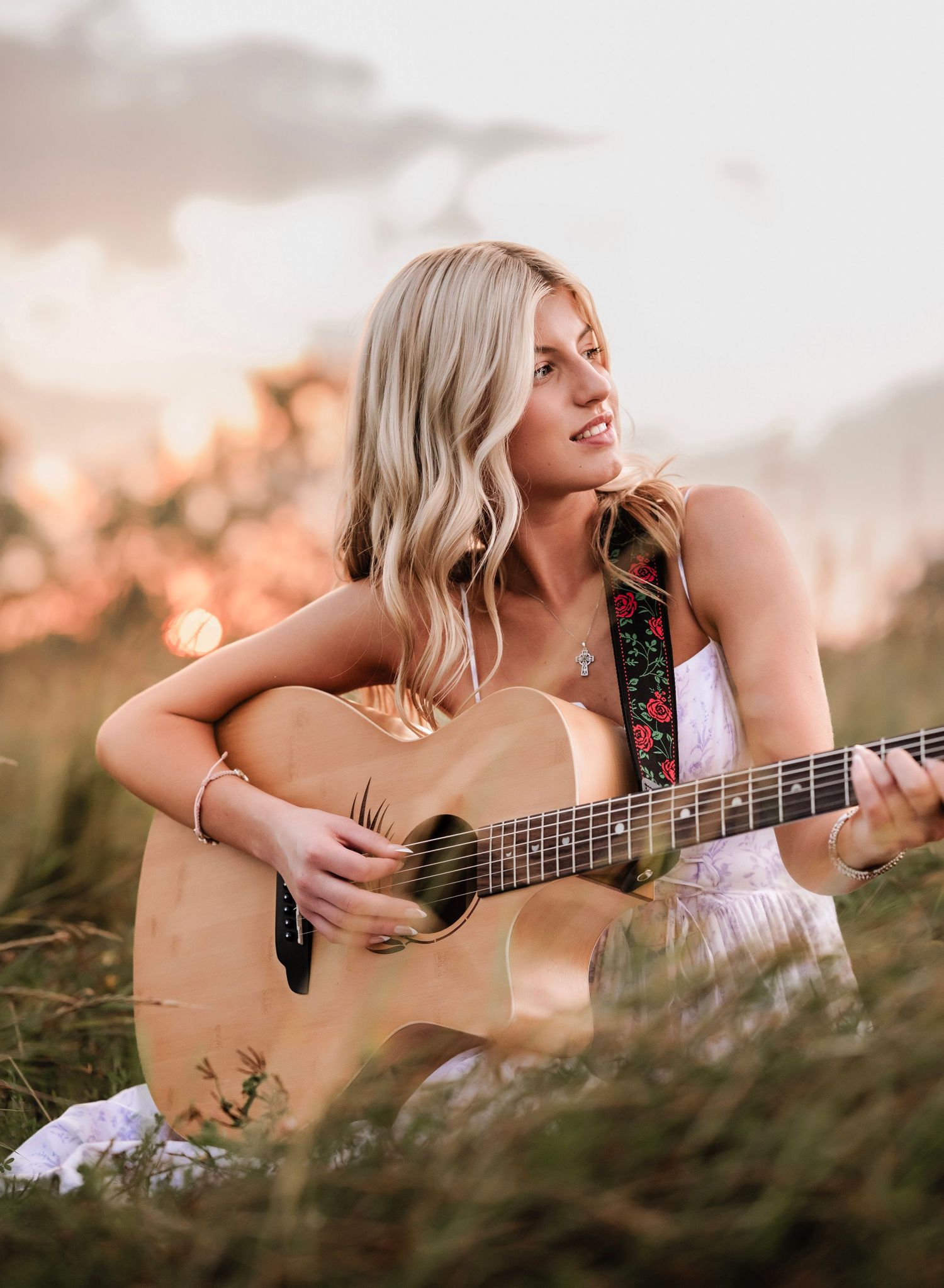 Acoustic guitar player in white dress sits in golden sunlit field during sunset while performing music outdoors.