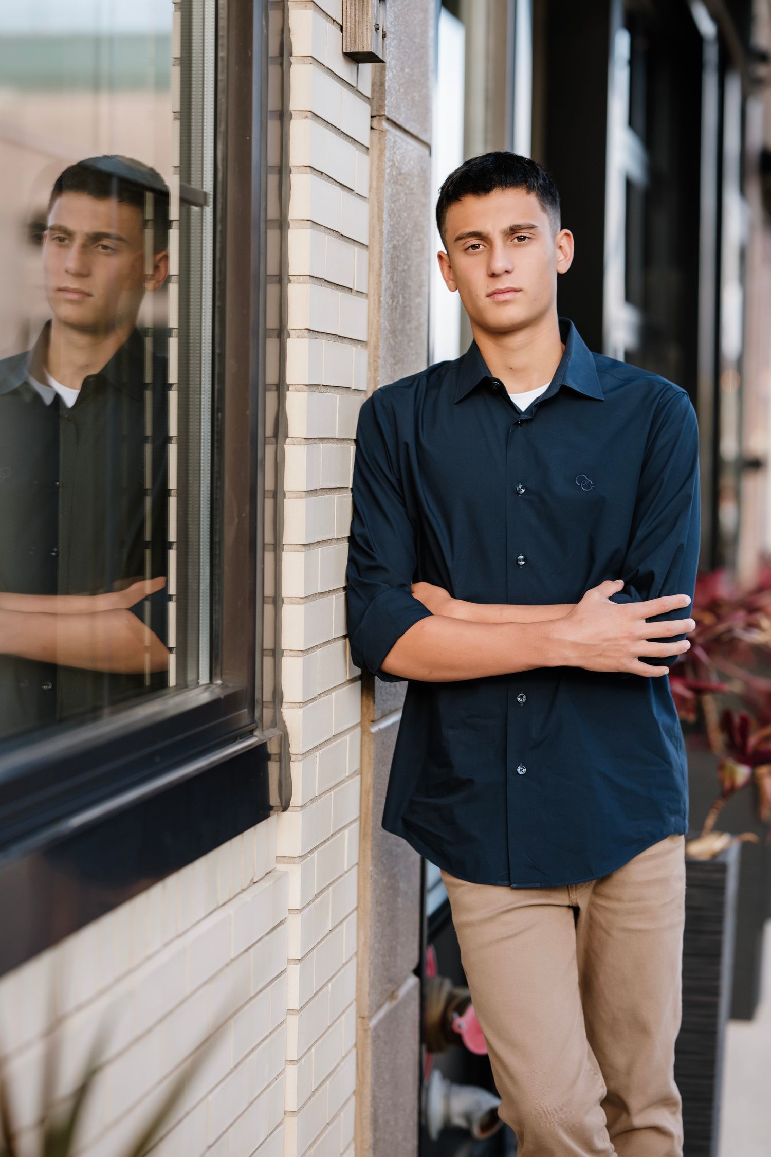 A person in a navy button-down shirt and khaki pants stands against a brick wall with their reflection visible in a window.