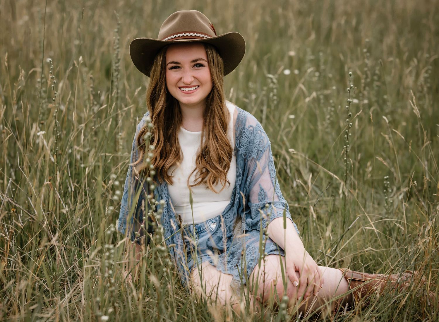 Person in brown cowboy hat and denim jacket sits in tall grass field during golden hour.
