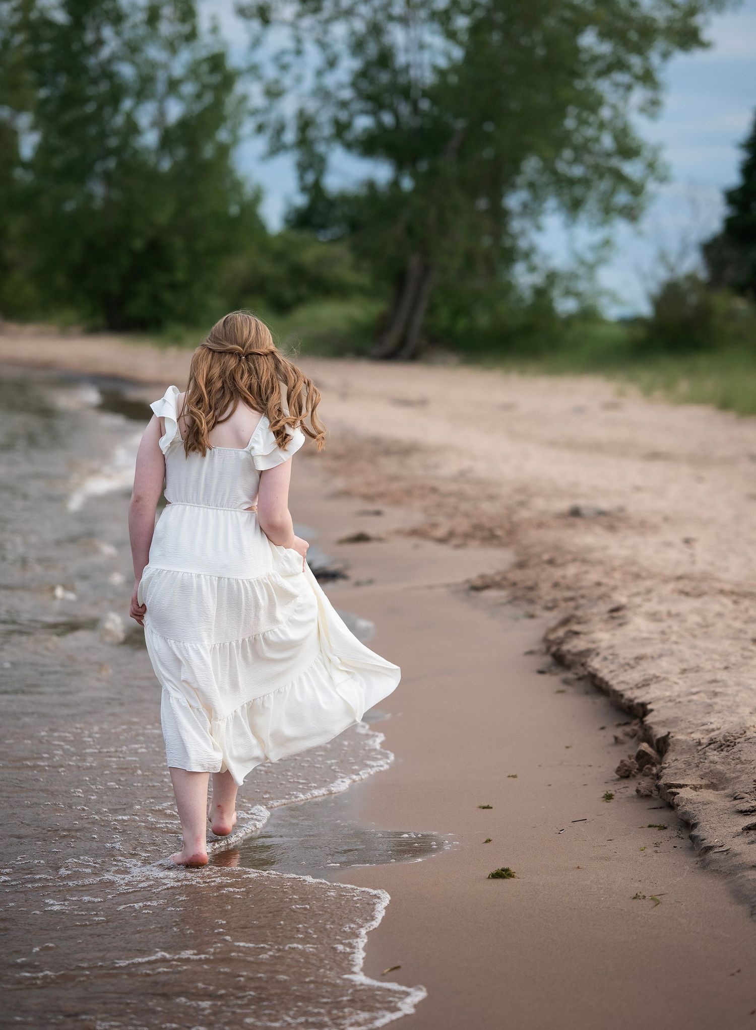 A small child in a white dress walks along the beach shoreline during a summer day.