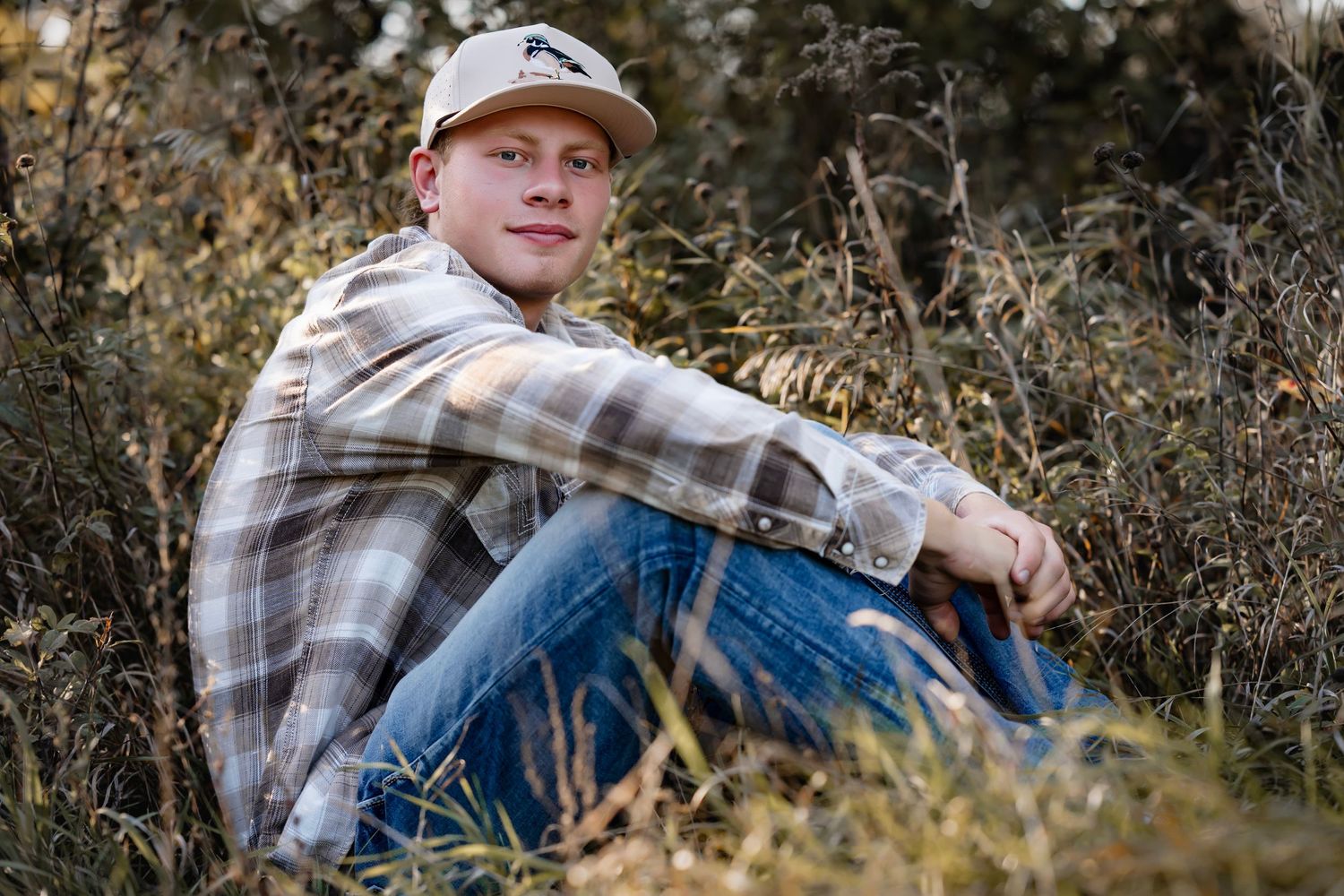 Person wearing plaid shirt and baseball cap sits outdoors in autumn setting with natural brush and foliage.