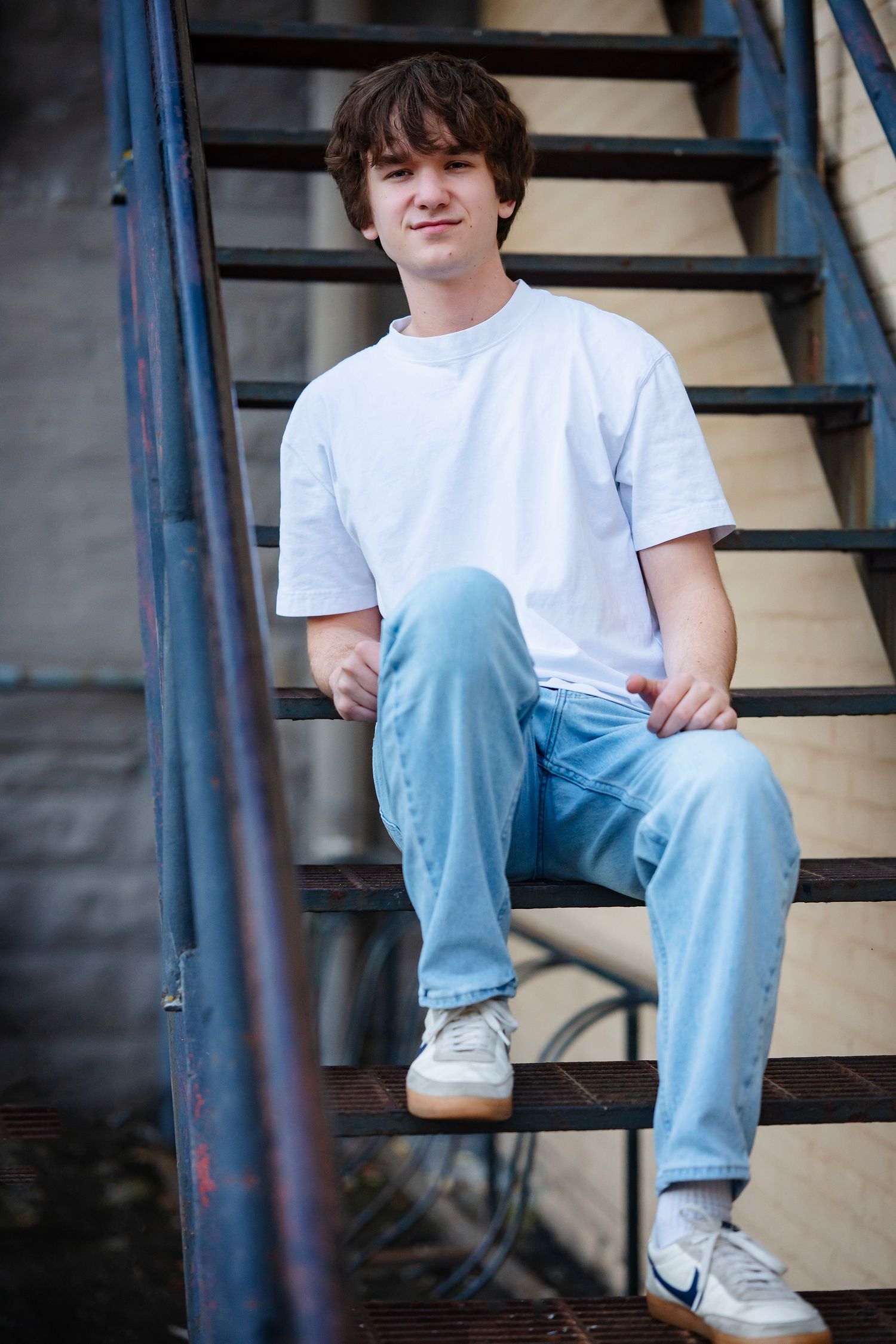 Person in white t-shirt and light blue jeans sitting casually on metal stairs wearing Nike sneakers.