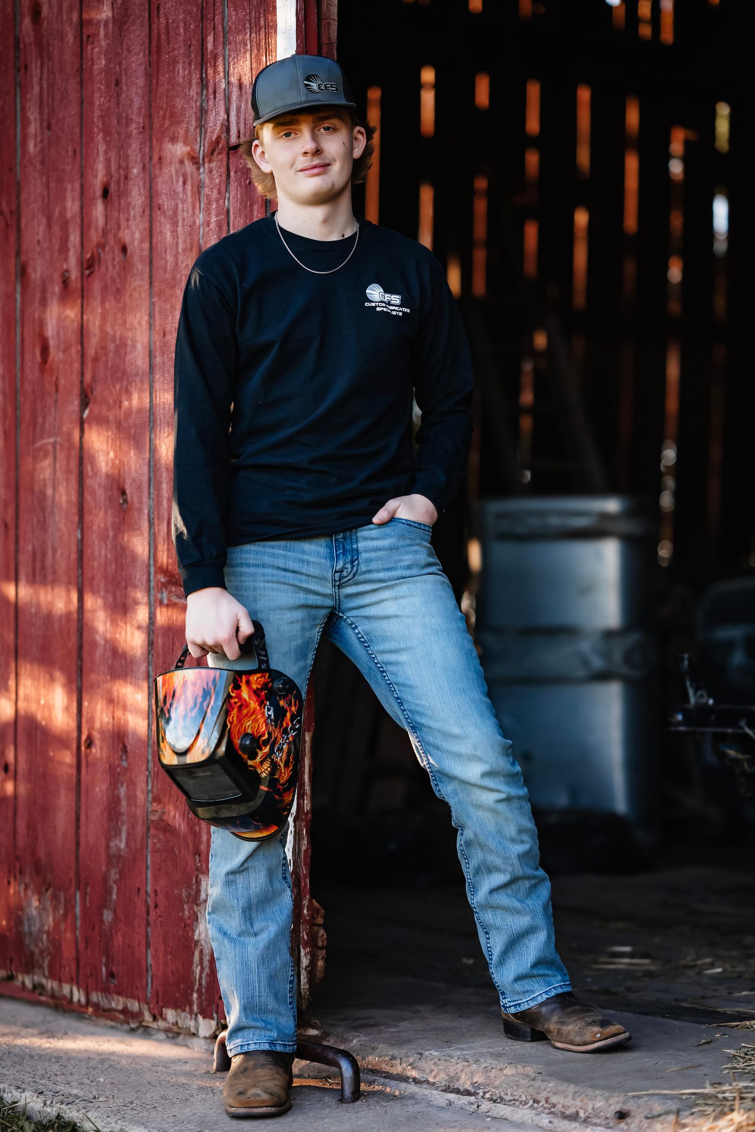Person wearing jeans and black shirt leans against rustic red barn door in casual outdoor portrait series.