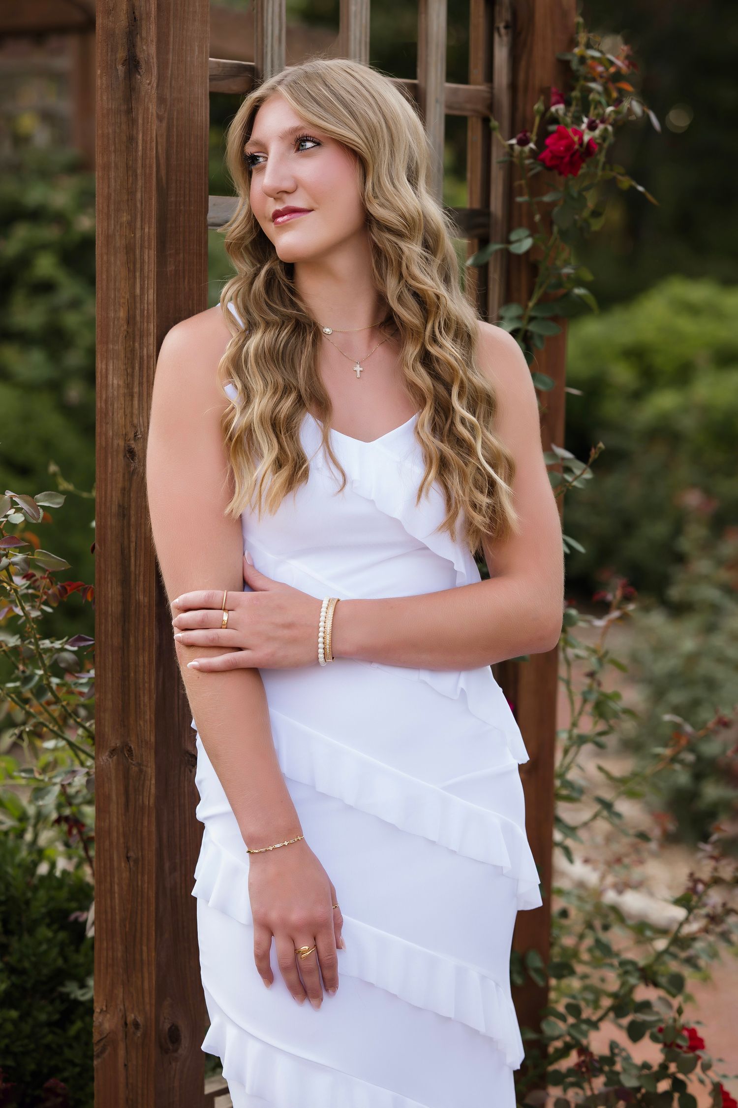 Young woman in white ruffled dress posing by wooden garden arbor with roses, long wavy blonde hair.