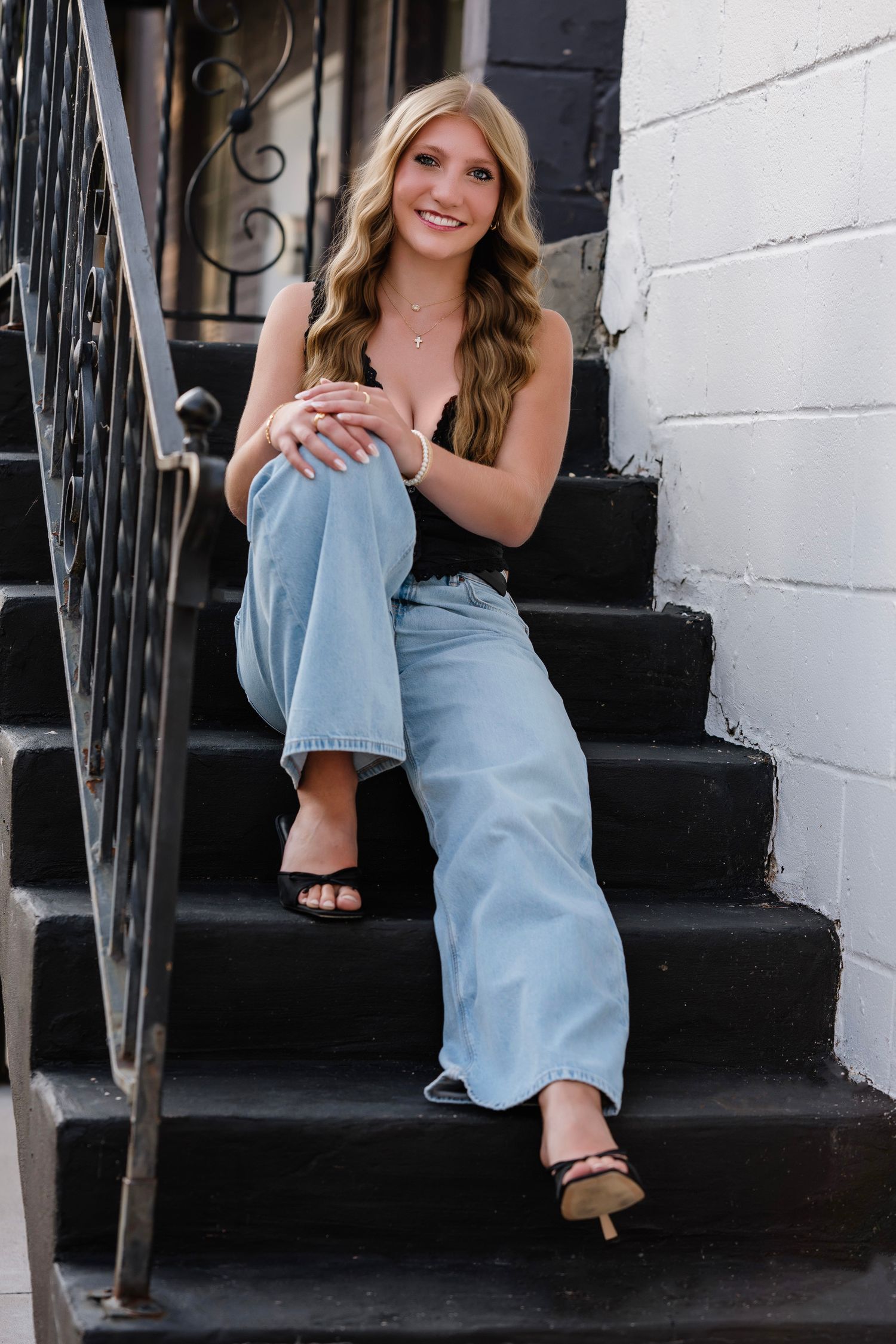 Young woman with blonde hair sits smiling on black stairs, wearing a black top and light blue wide-leg jeans.