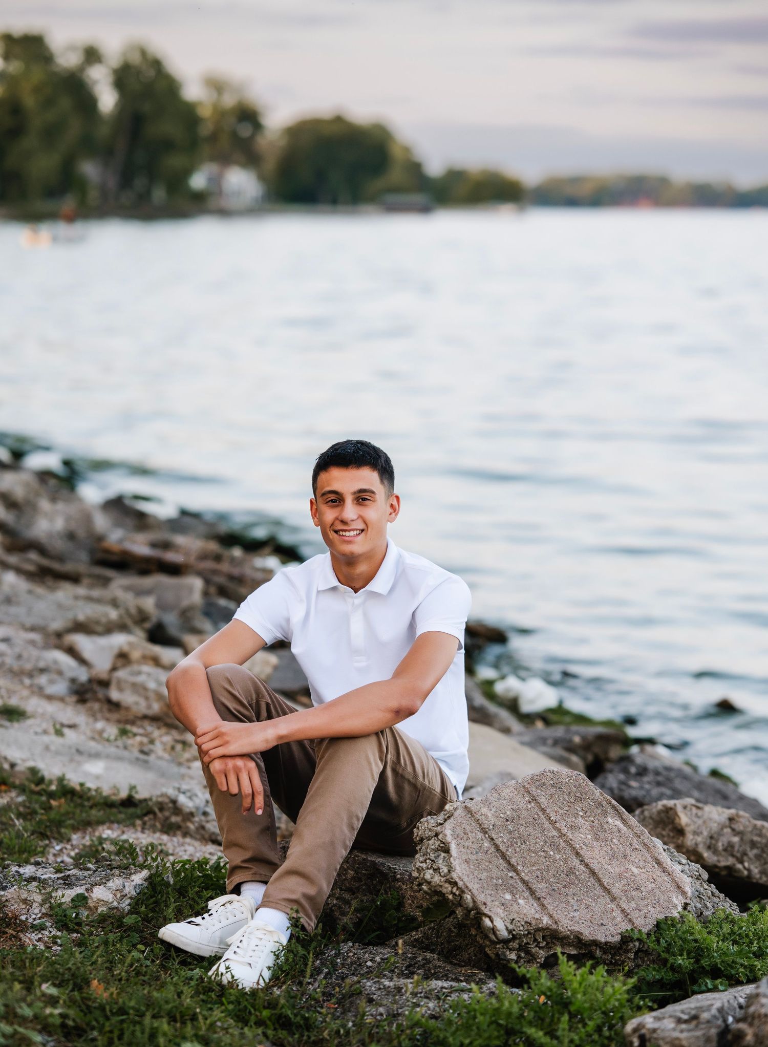 Young man in white polo and khaki pants sitting on rocks by a lake shore at sunset.