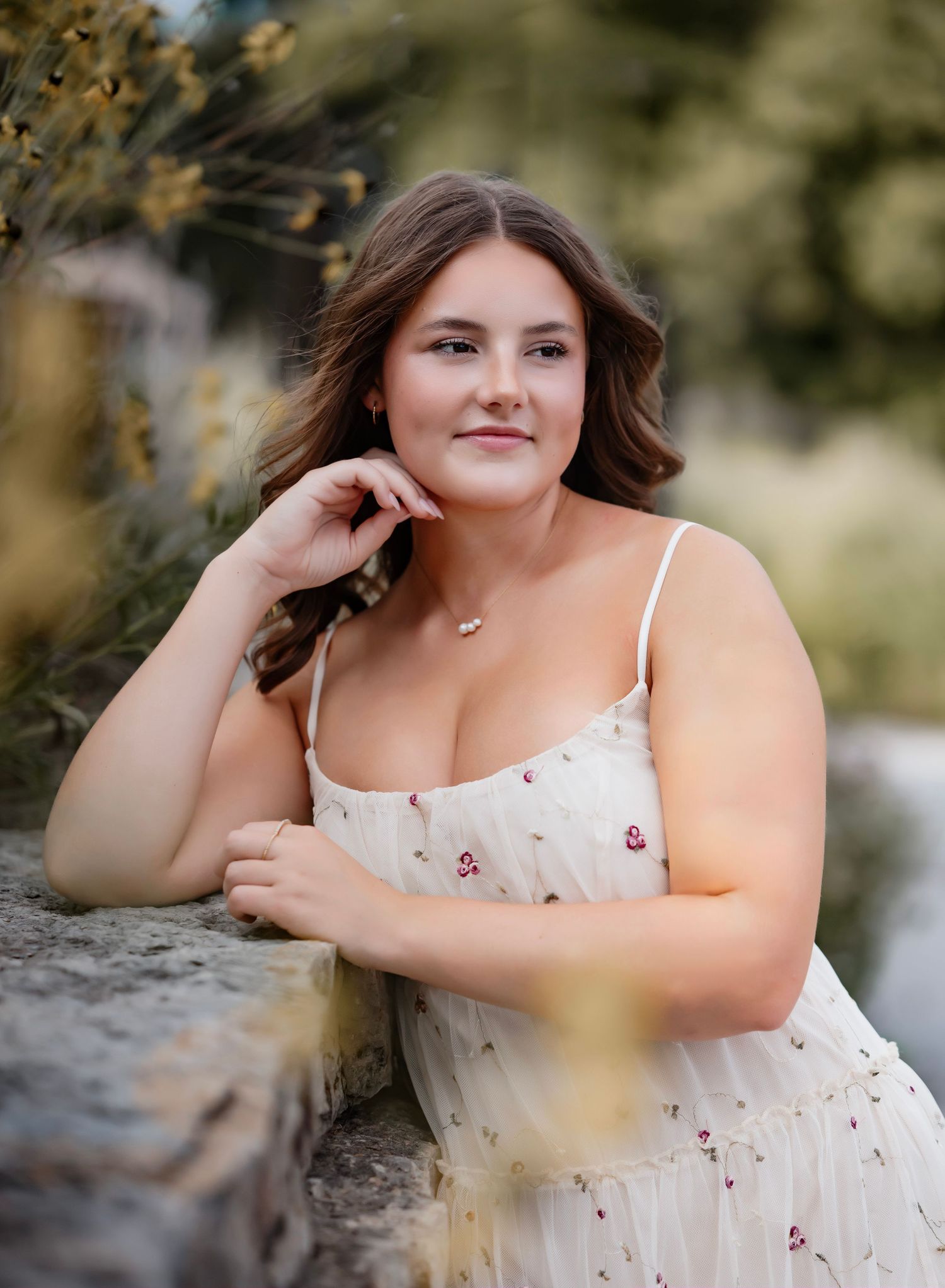 A person in a white floral sundress poses outdoors against a blurred natural background on a sunny day.