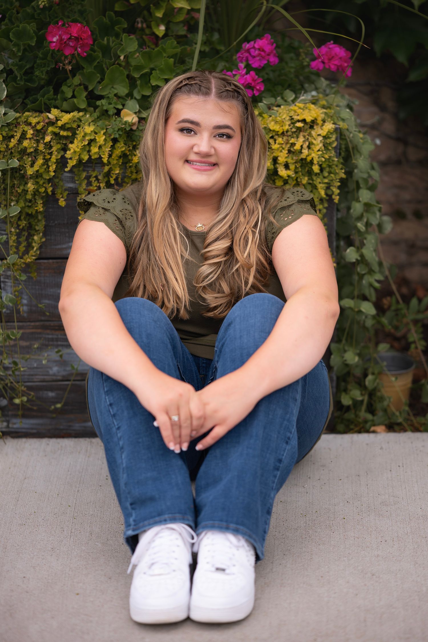 Young woman with blonde hair sits smiling on steps, wearing jeans and olive top, with colorful flowers behind her.