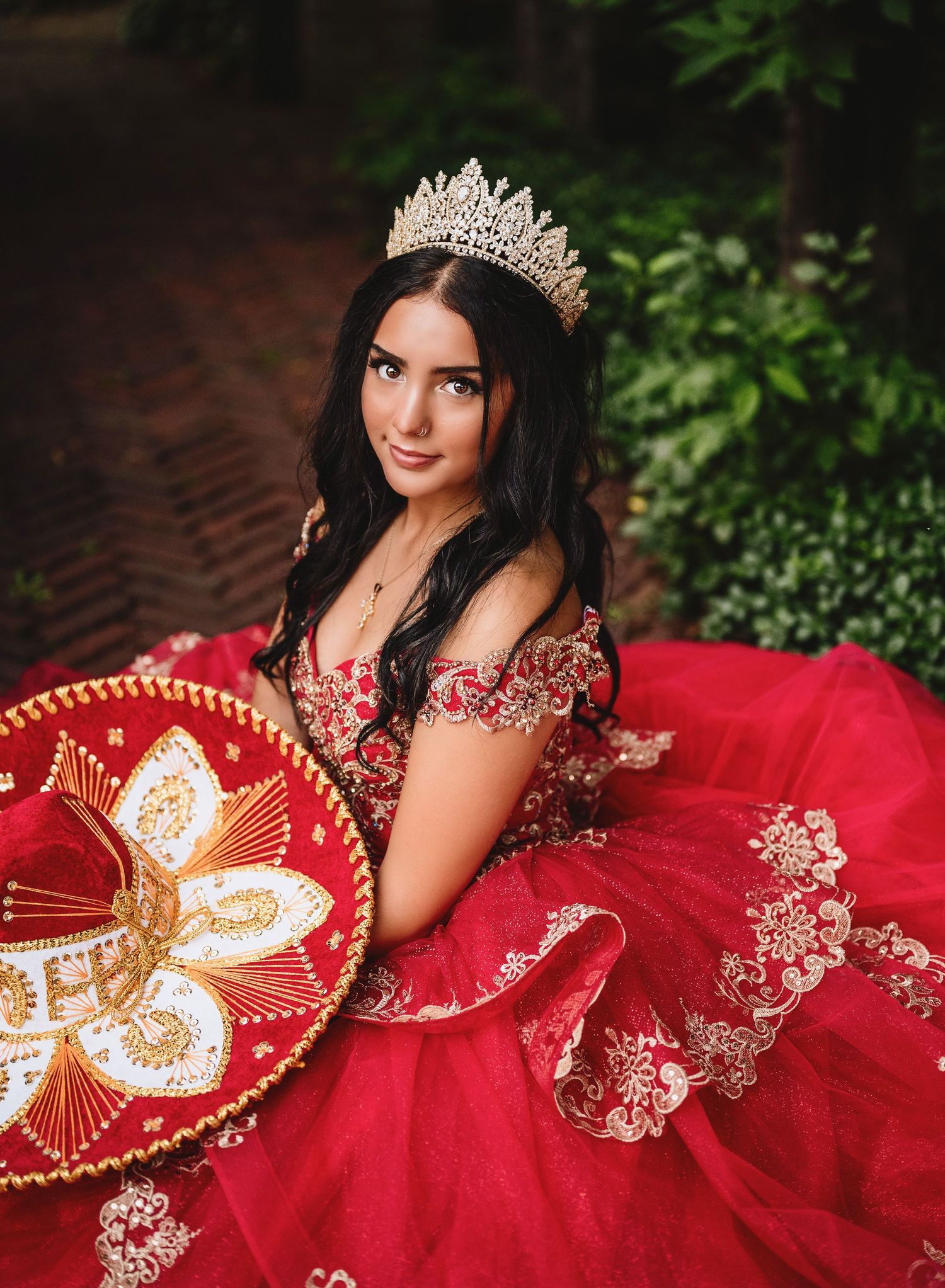 Elegant quinceañera portrait in vibrant red with gold embroidery details, paired with a crystal crown and decorative sombrero.
