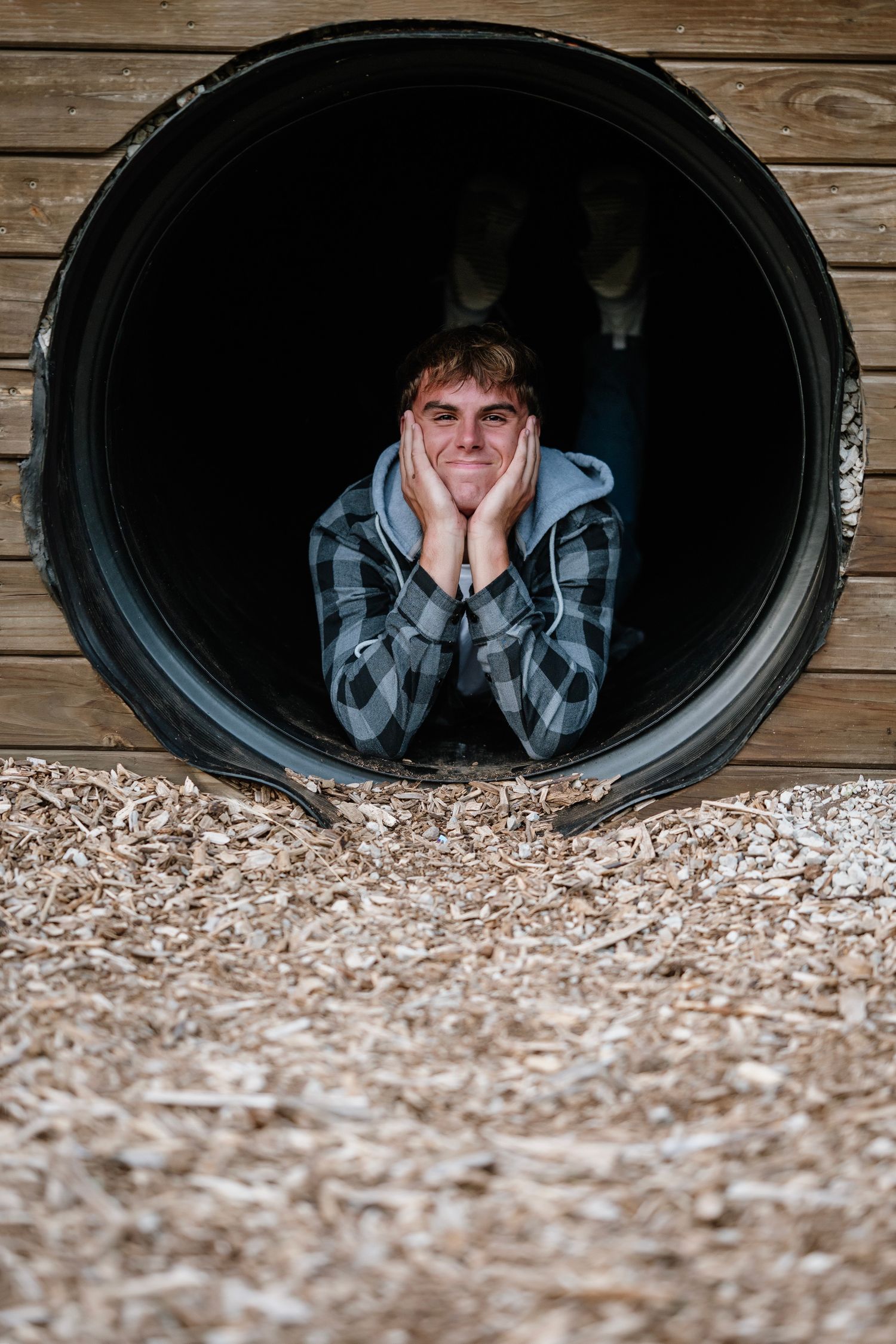 A person peeks through a dark circular opening in a wooden wall with wood shavings scattered below.