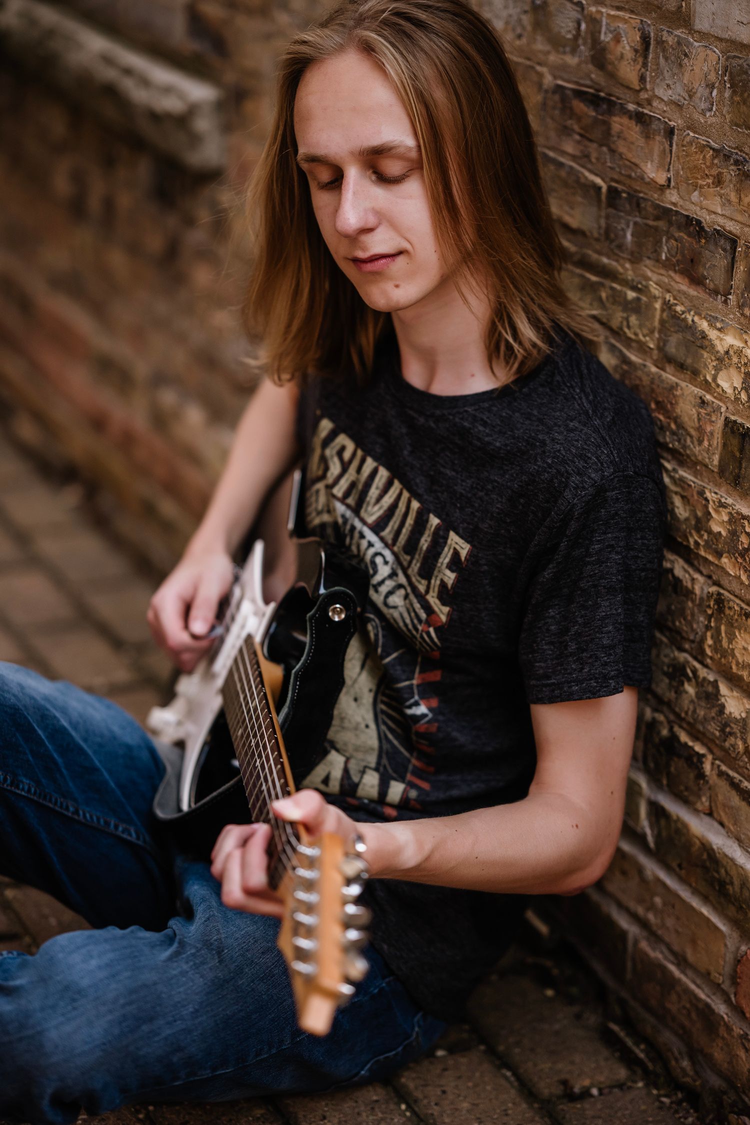 Young man with long hair playing electric guitar while sitting against a brick wall wearing a Nashville t-shirt.