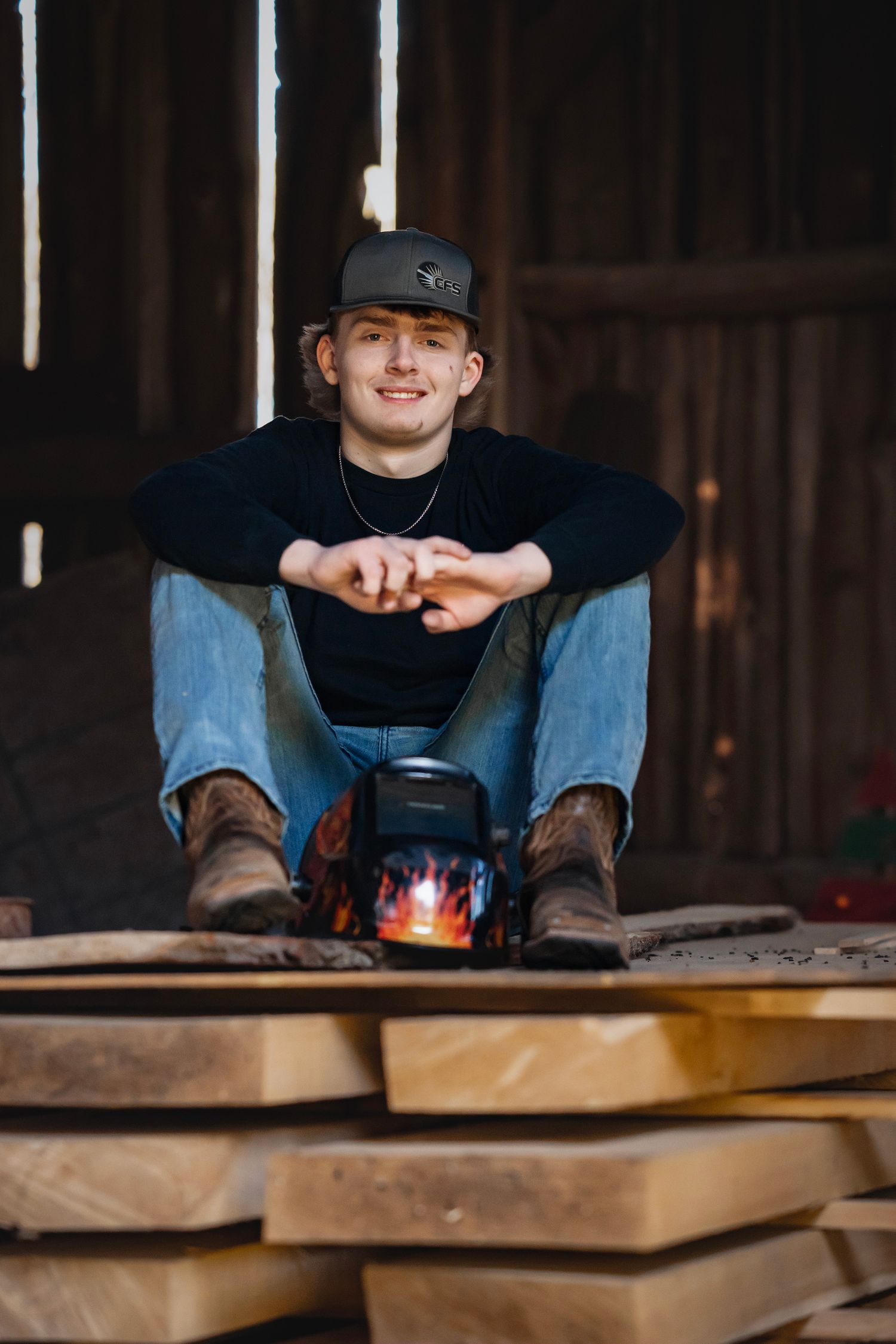 Person wearing a baseball cap sits at wooden table with stacks of lumber in a dimly lit workshop setting.