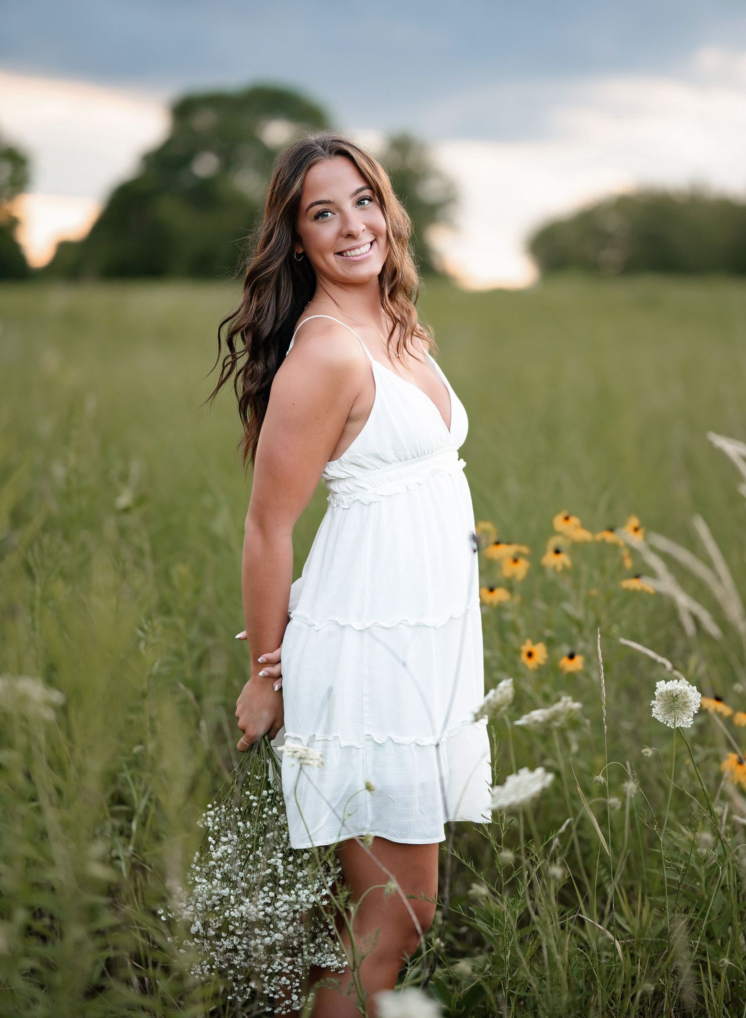 A young woman in a white sundress smiles while standing among wildflowers in a lush green meadow at sunset.