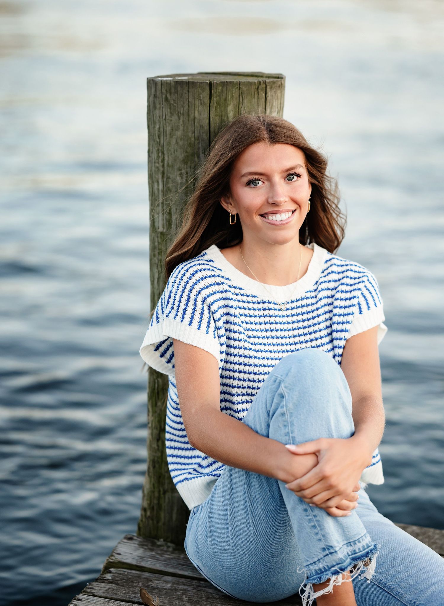 Smiling young woman in blue striped top and jeans sitting on a wooden dock by the water.