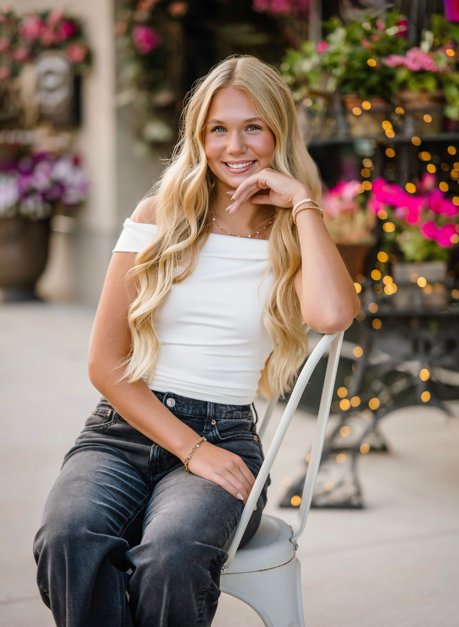 A person in a white off-shoulder top and dark jeans poses outdoors near floral decorations and twinkle lights.