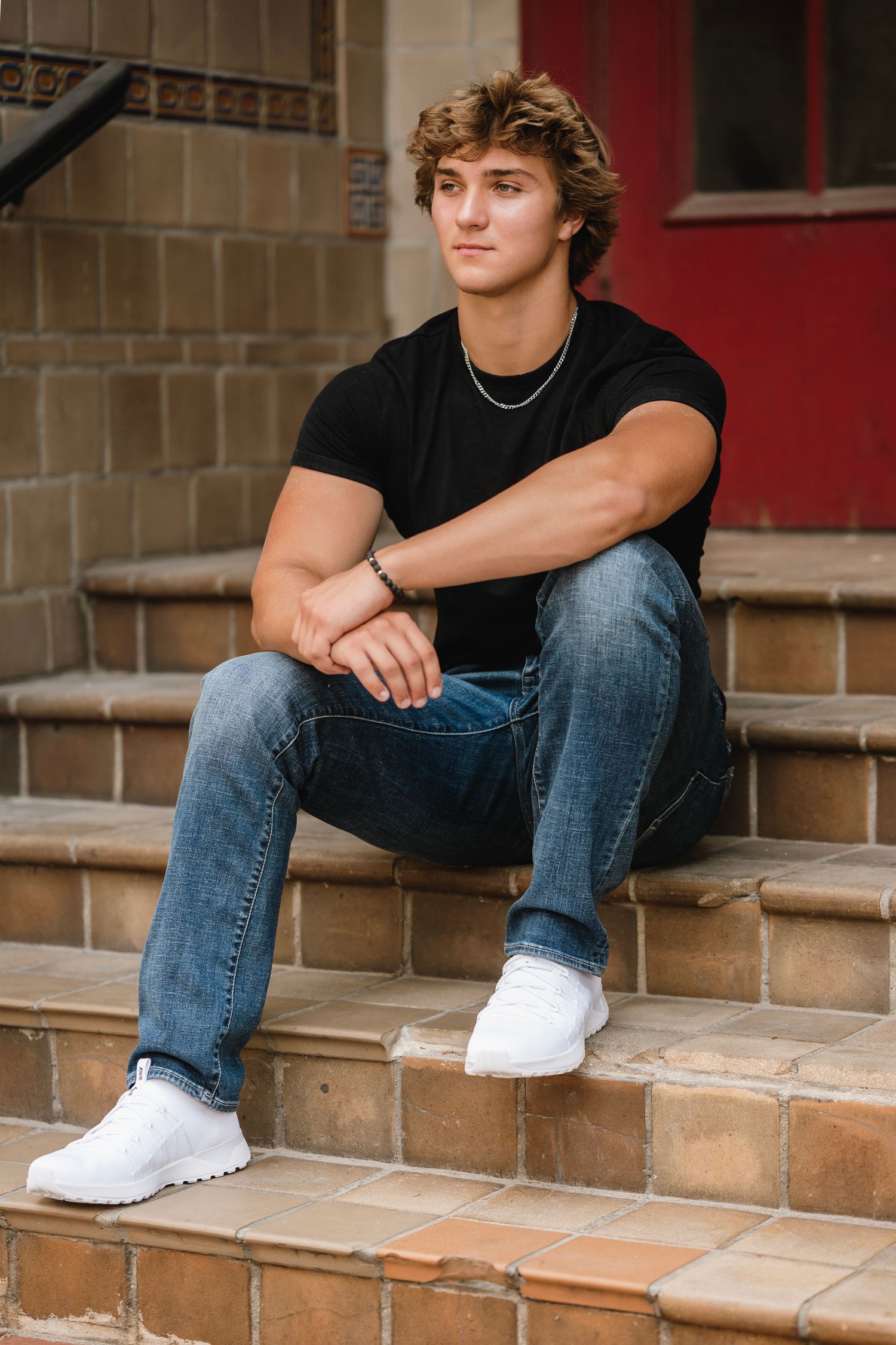 Person in jeans and black shirt sits casually on tan stone steps wearing white shoes.