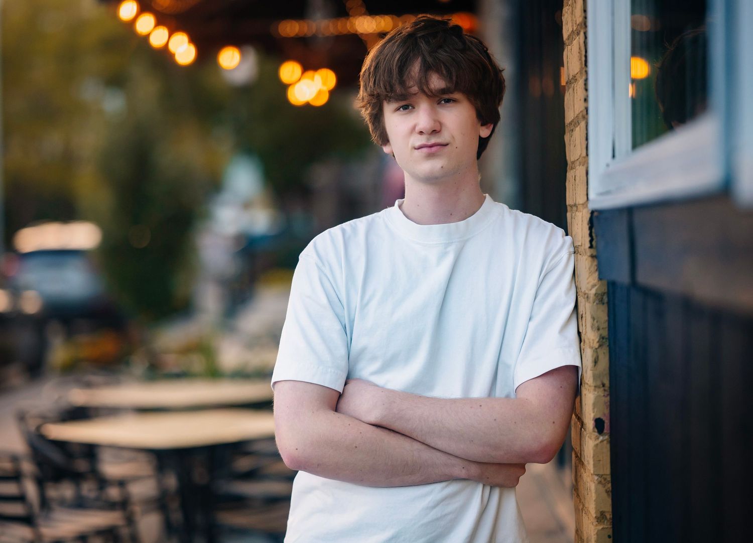 Person in white t-shirt poses against urban backdrop with bokeh lights and greenery in background.