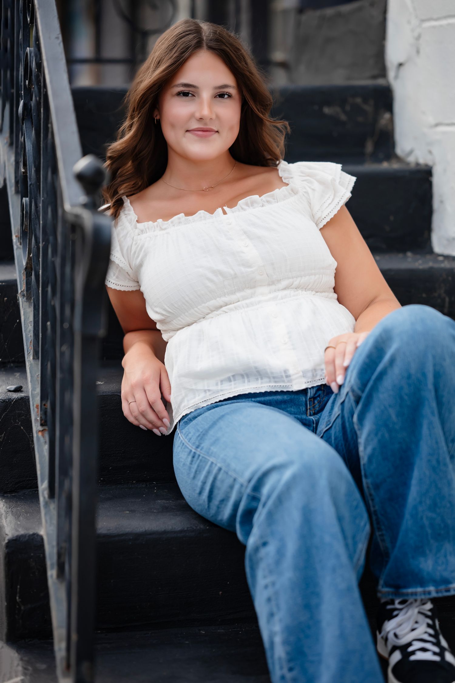 A person in a white off-shoulder top and blue jeans sits casually on outdoor concrete steps.