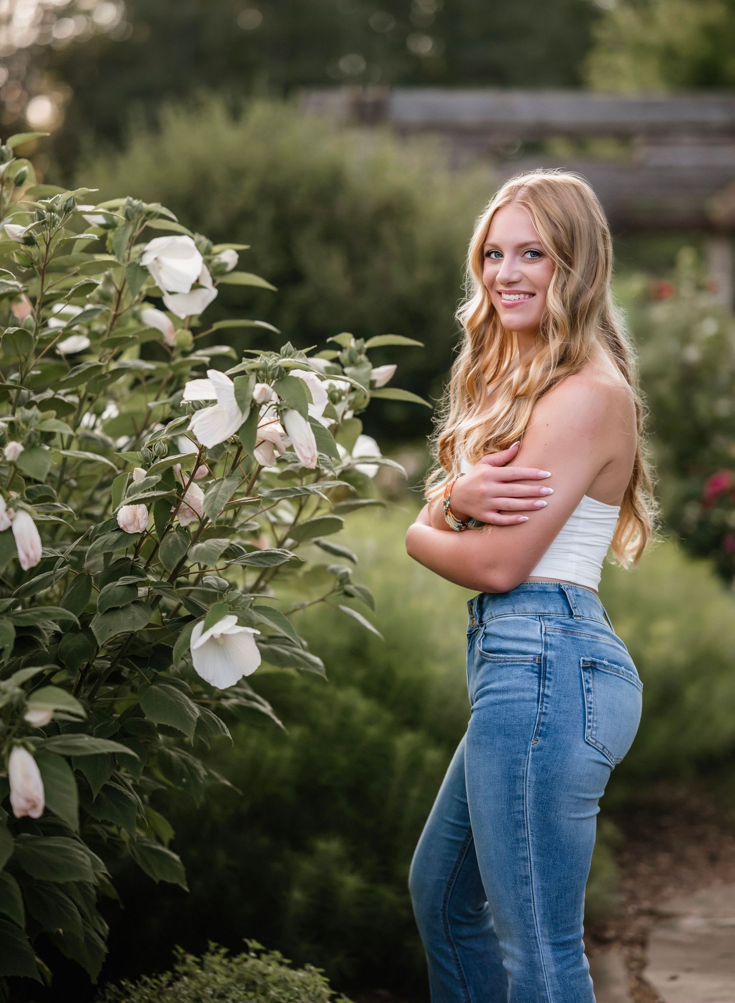 A woman in blue jeans stands among blooming white magnolia flowers in a lush garden setting.