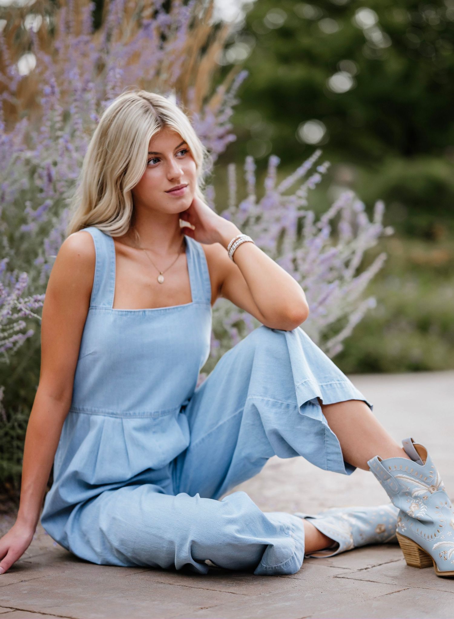 A woman in a light blue summer dress reclines on stone steps near blooming lavender flowers in an outdoor garden setting.