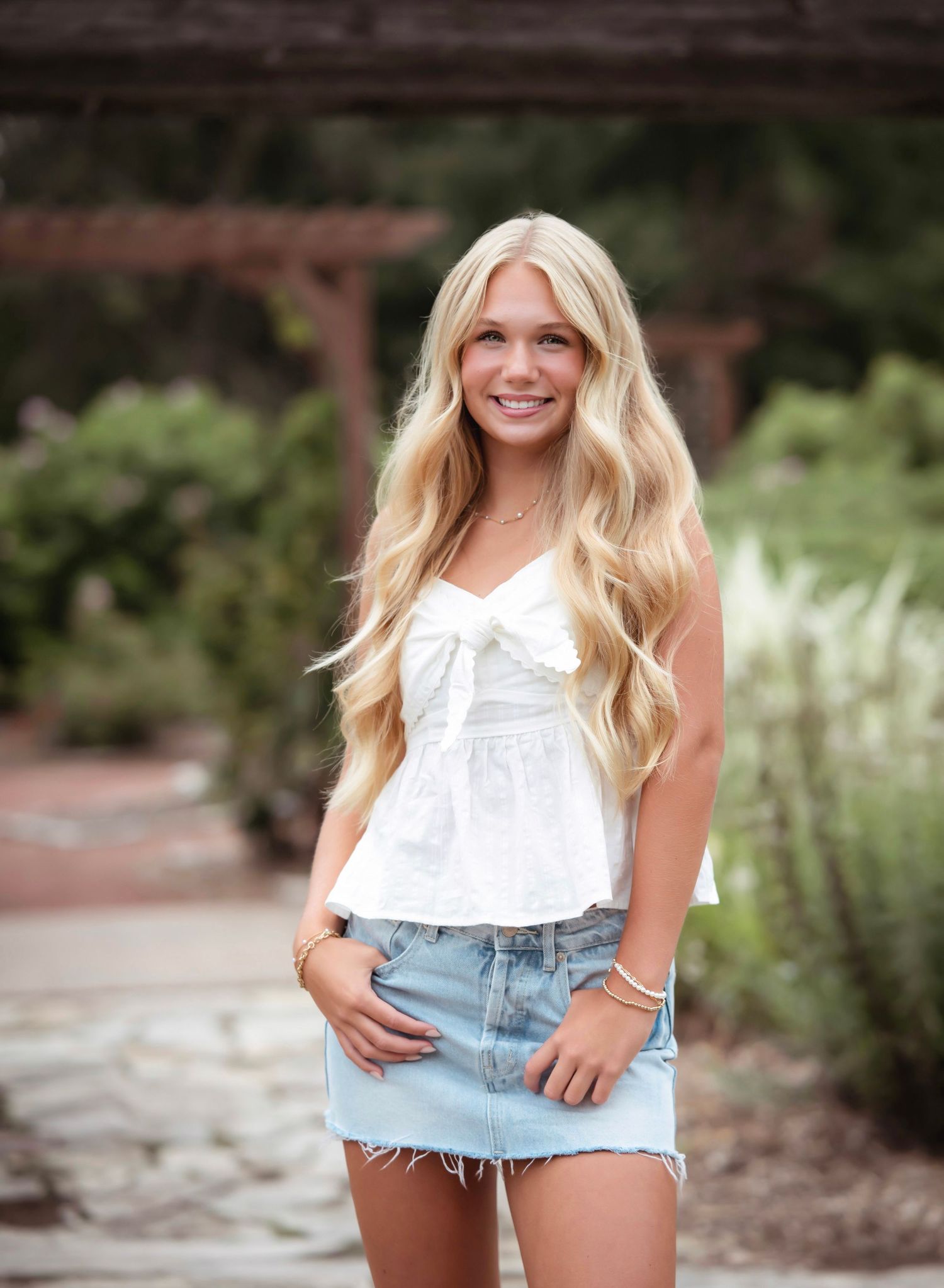 Young woman in white sleeveless top and denim mini skirt poses outdoors with long wavy blonde hair.